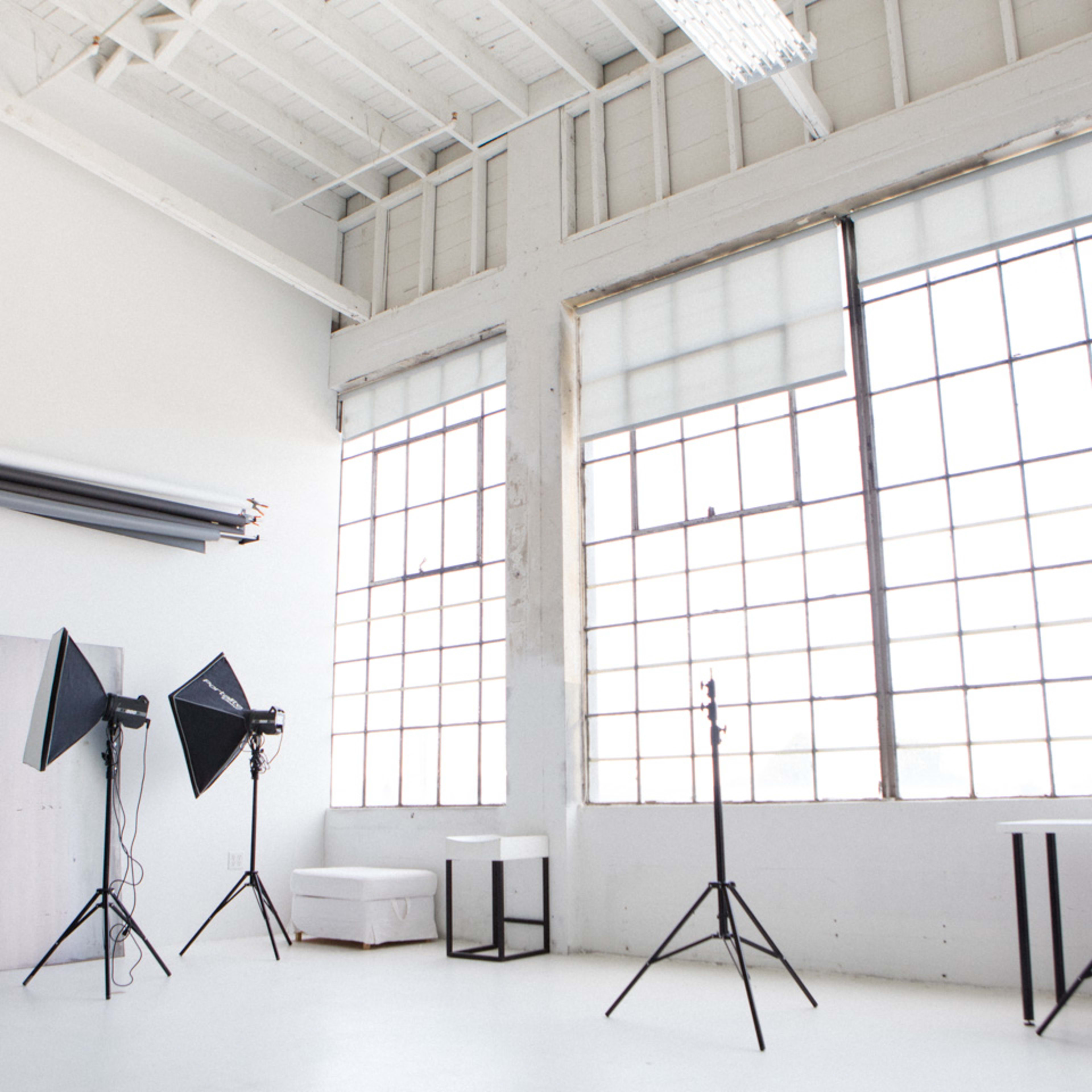 A spacious photo studio featuring large windows, backdrop rolls, and lighting equipment set up on stands.