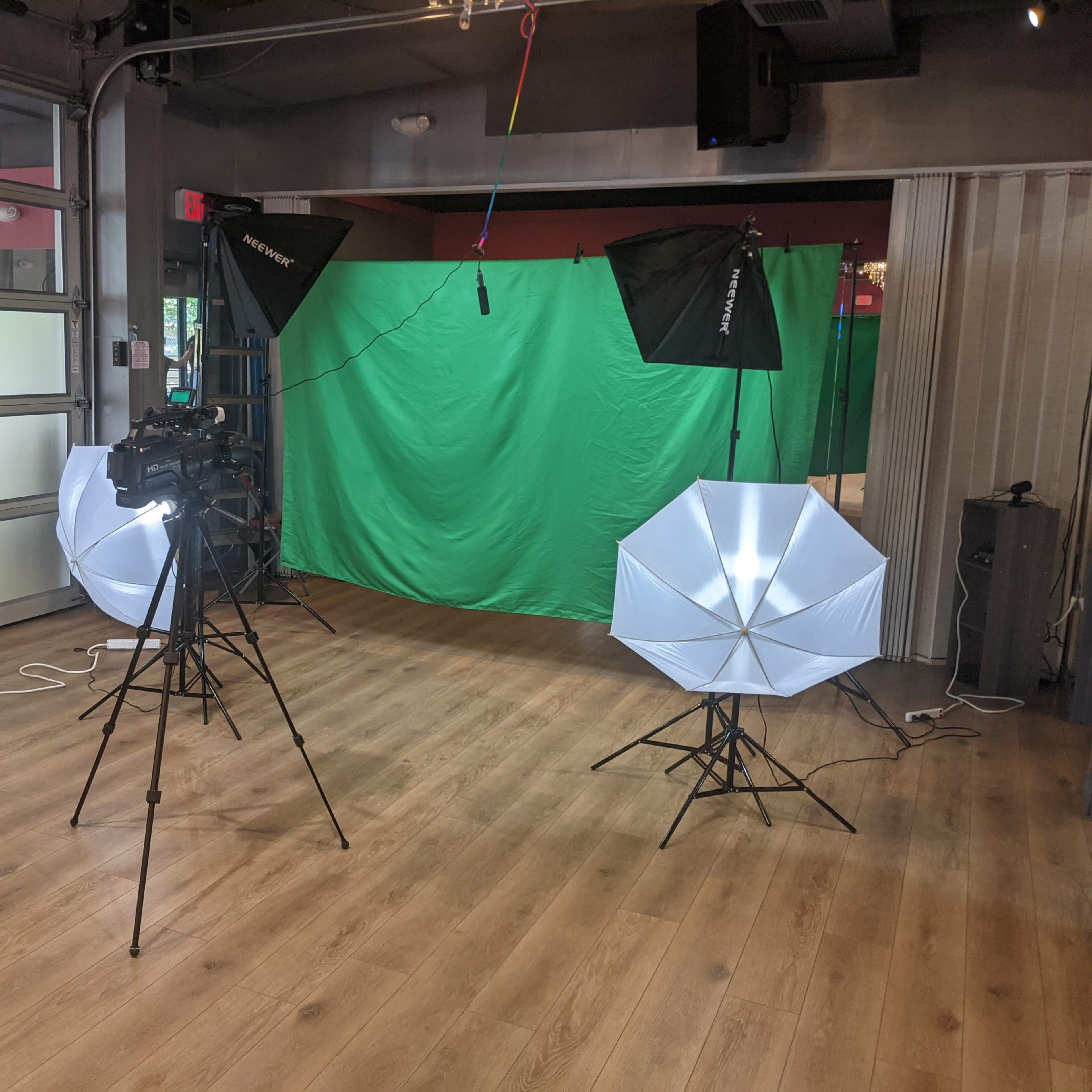 A studio setup with two softbox lights, a camera on a tripod, and a green screen in a room with wooden flooring.