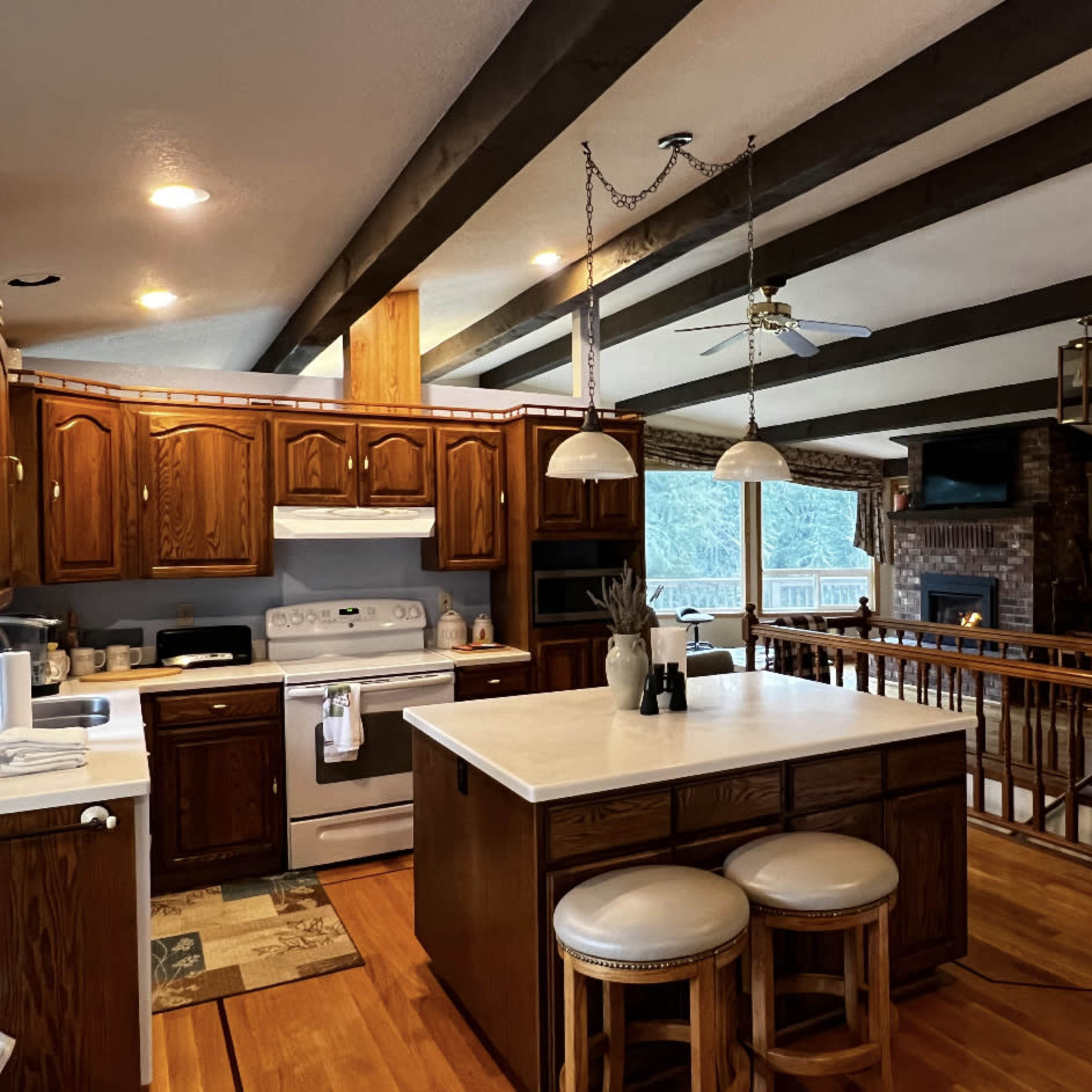 A spacious kitchen with wooden cabinets, a central island with bar stools, and a view of a living area featuring a fireplace and large windows.