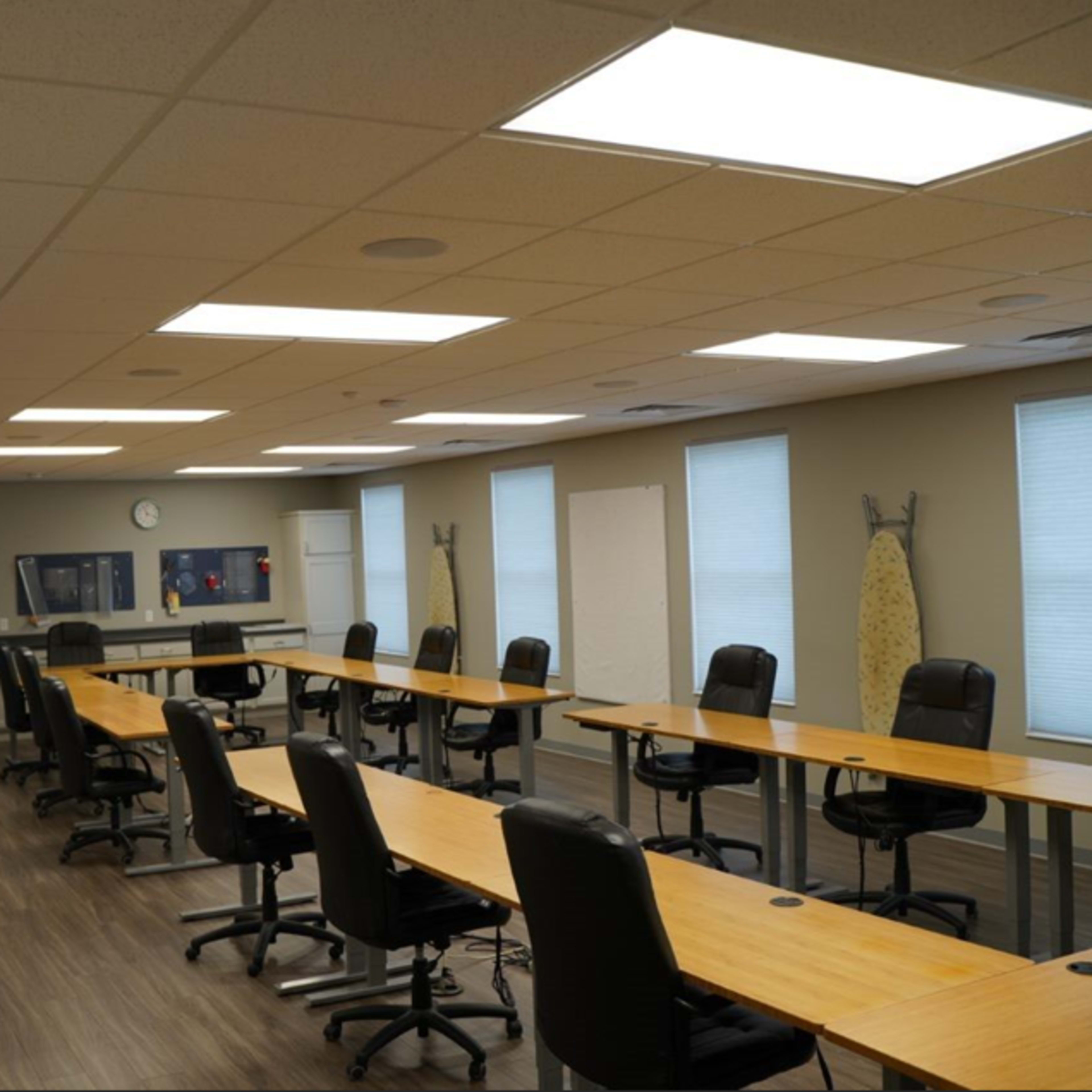 A well-lit classroom features several long tables and black rolling chairs arranged in a spacious layout, with whiteboards and windows along the walls.