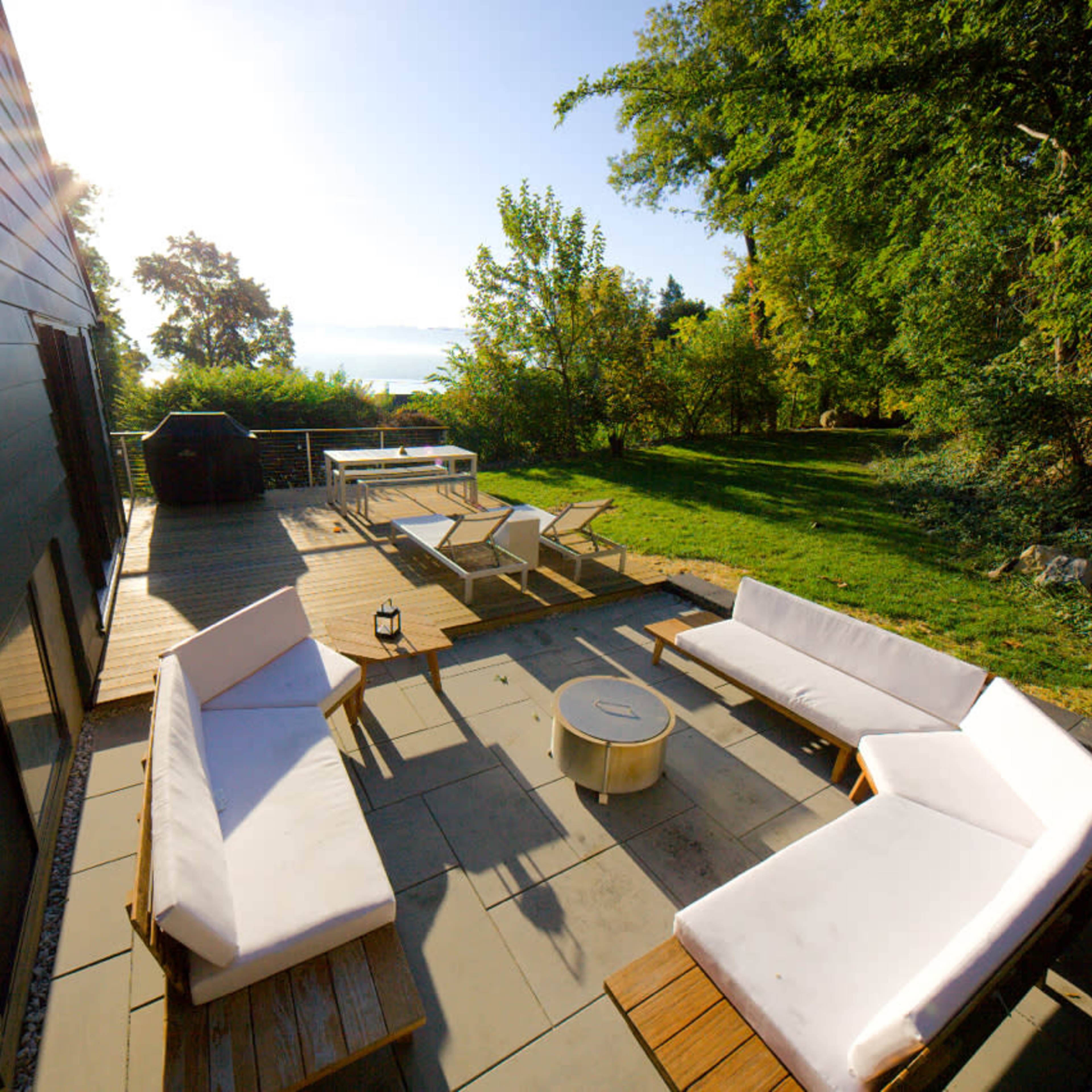 An outdoor seating area with several white couches and lounge chairs on a wooden deck surrounded by greenery.