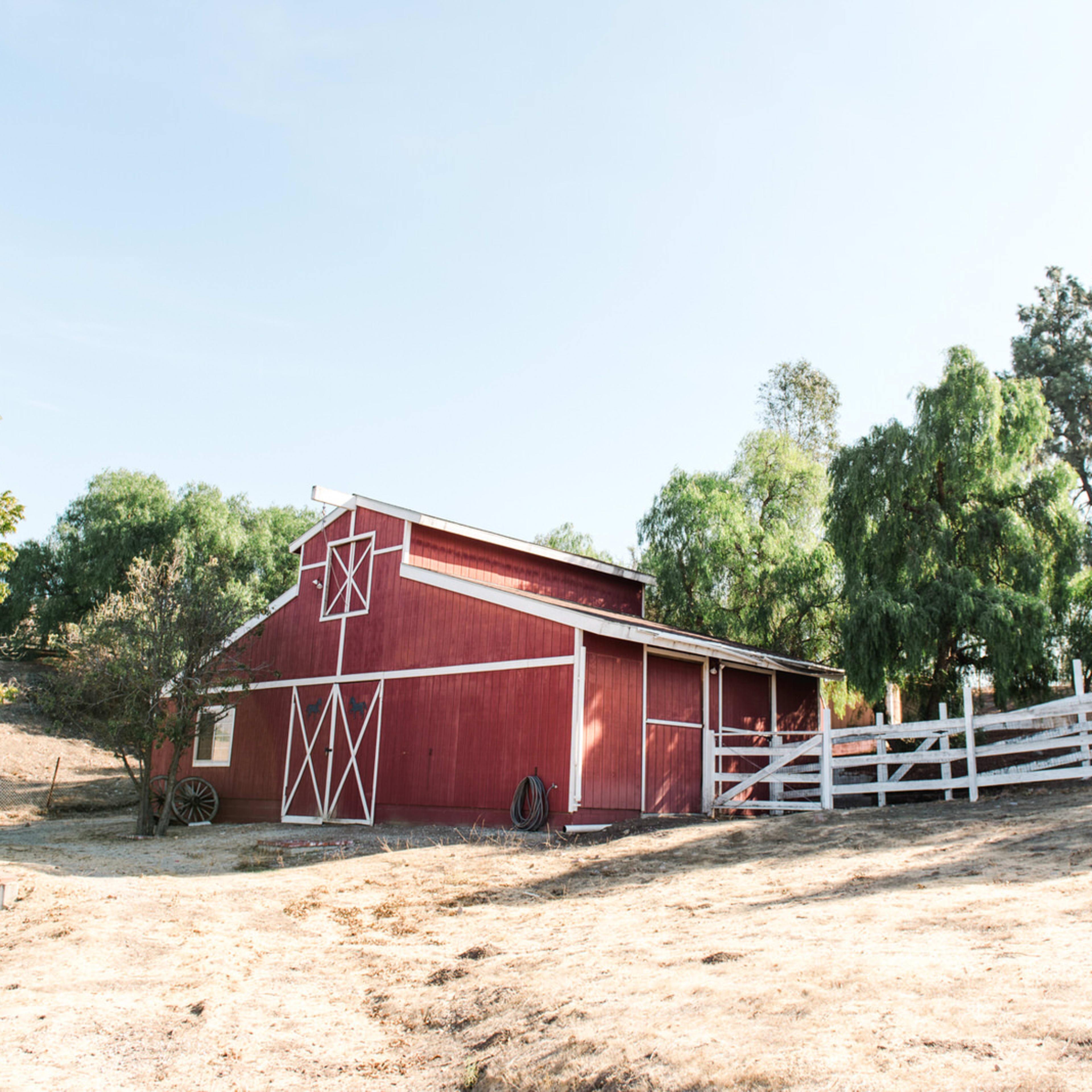 A red barn with white trim stands on a hillside surrounded by trees and a white fence.