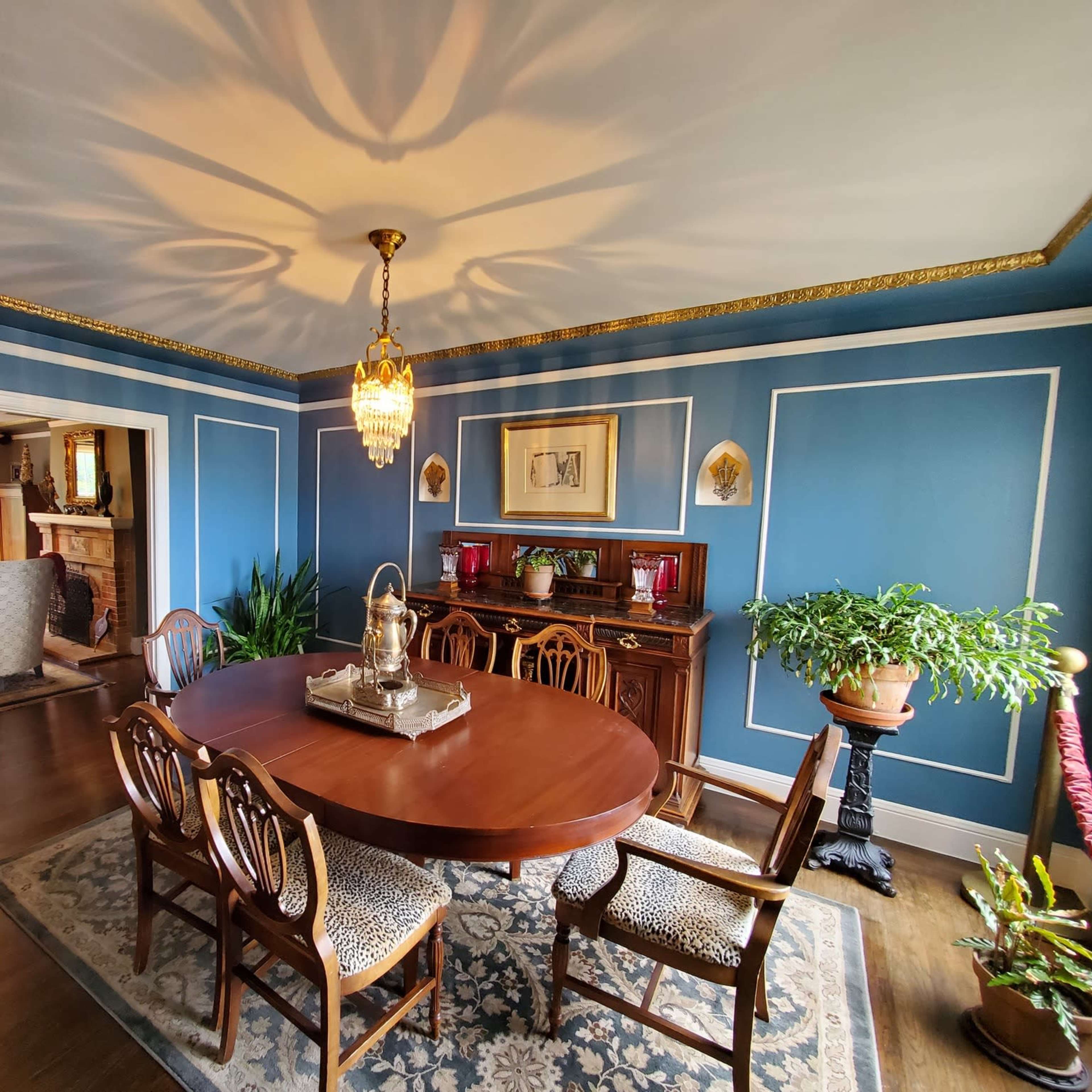 A dining room with a wooden table surrounded by chairs, a chandelier hanging from the ceiling, and blue walls adorned with decorative moldings and plants.
