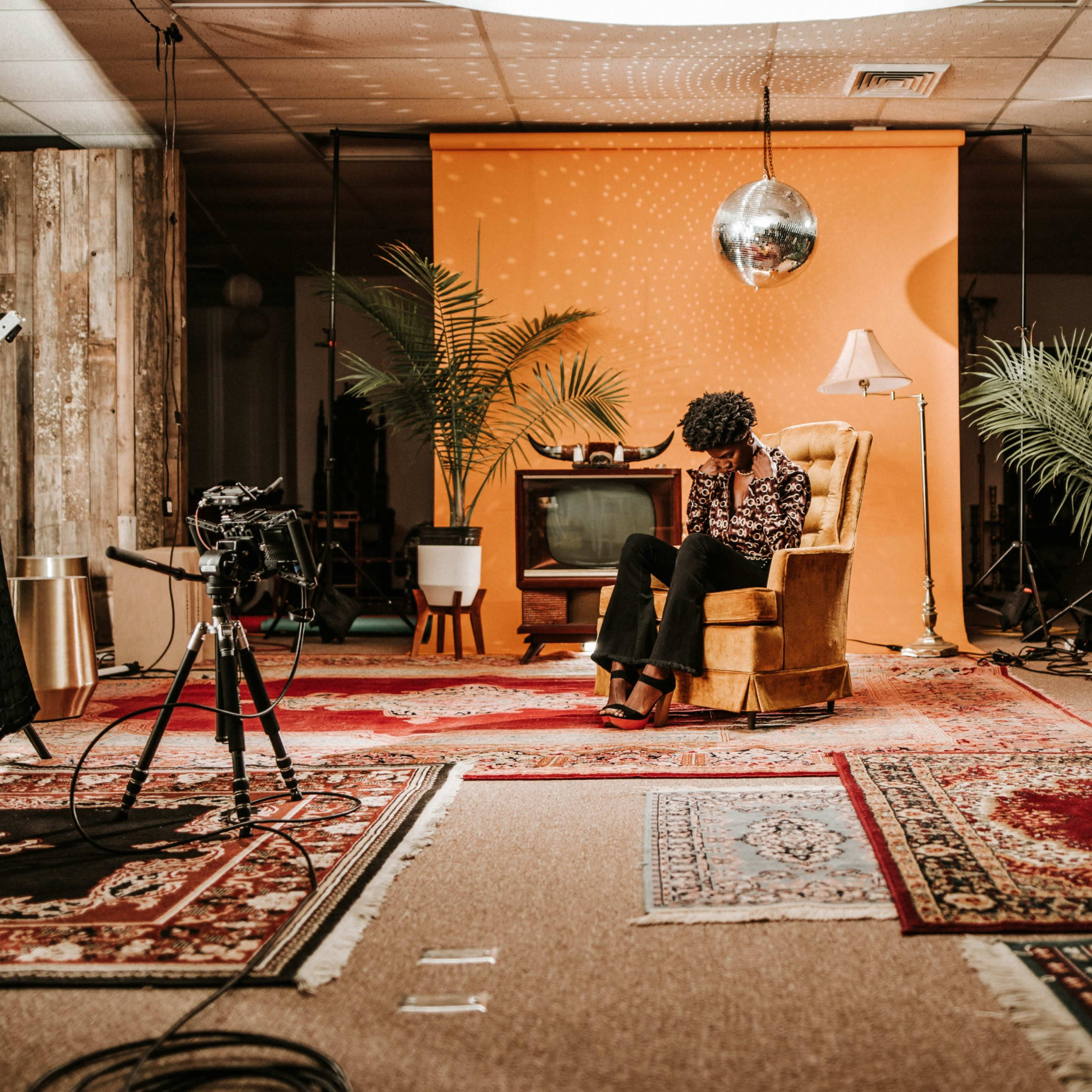 A person sits in a vintage armchair in a studio setting, surrounded by plants, a retro television, and camera equipment on a patterned rug.