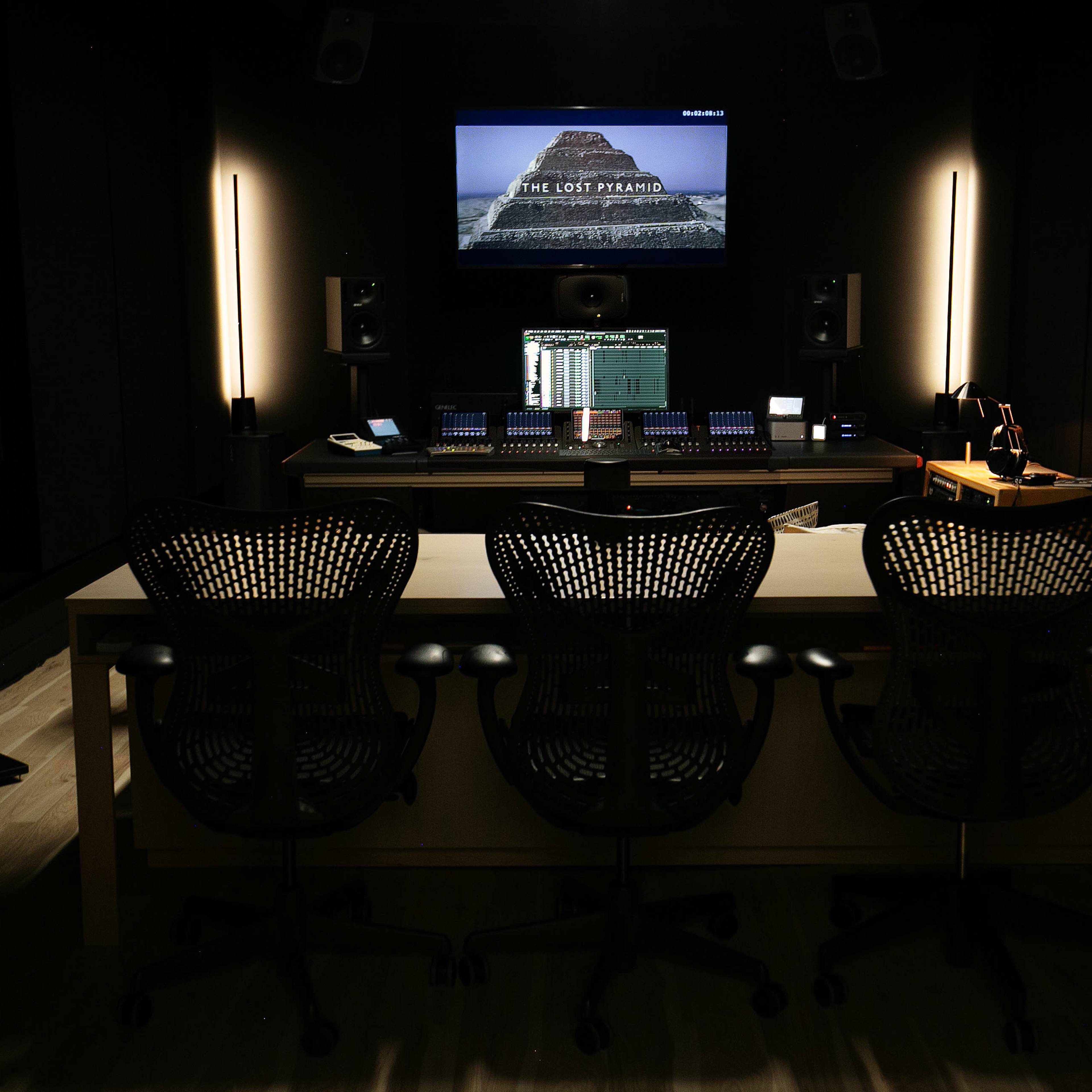A dark recording studio with a central desk featuring audio equipment, three black office chairs, and a screen displaying "THE LOST PYRAMID."