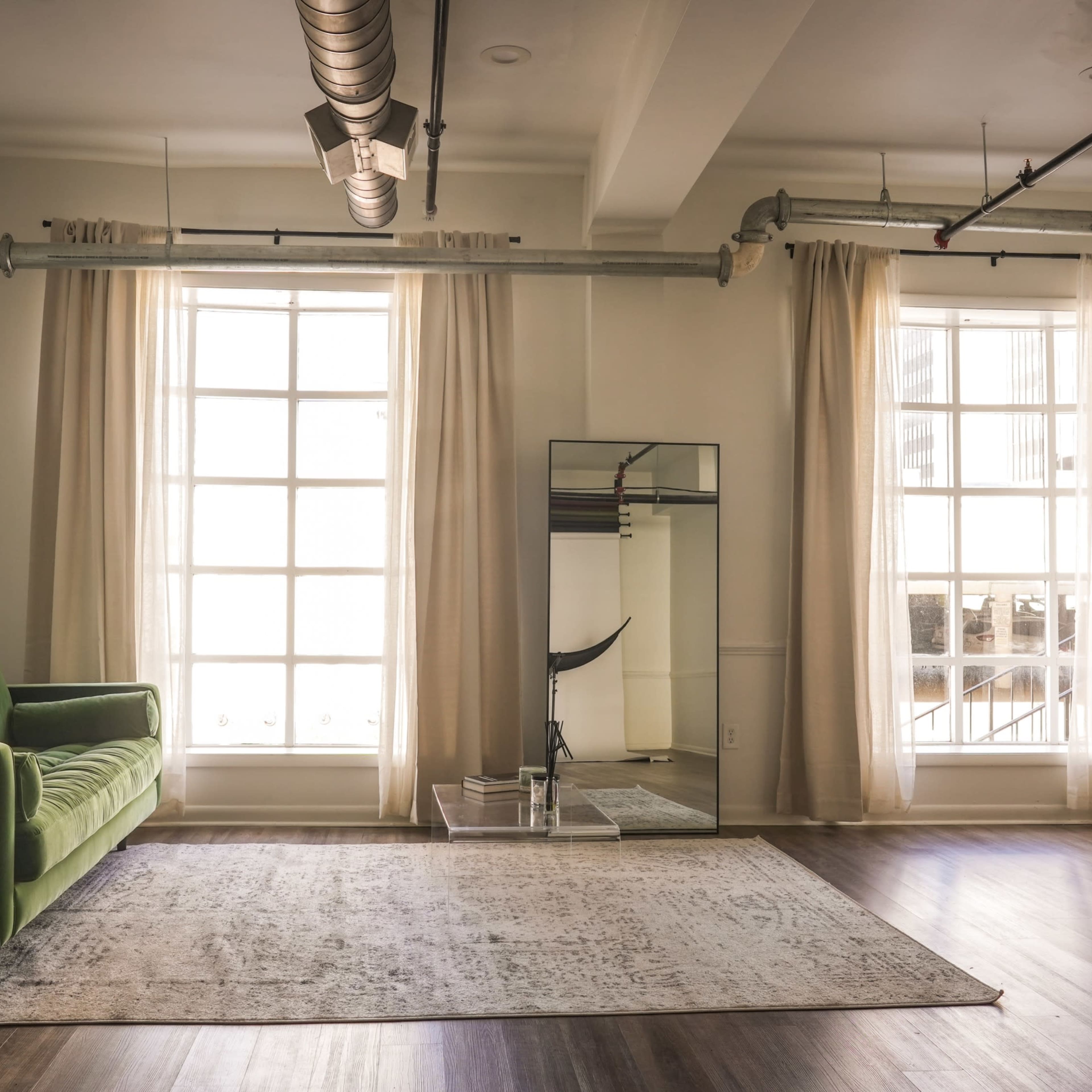 A bright room features a green sofa, large windows with sheer curtains, a mirror, and a patterned rug on a wooden floor.