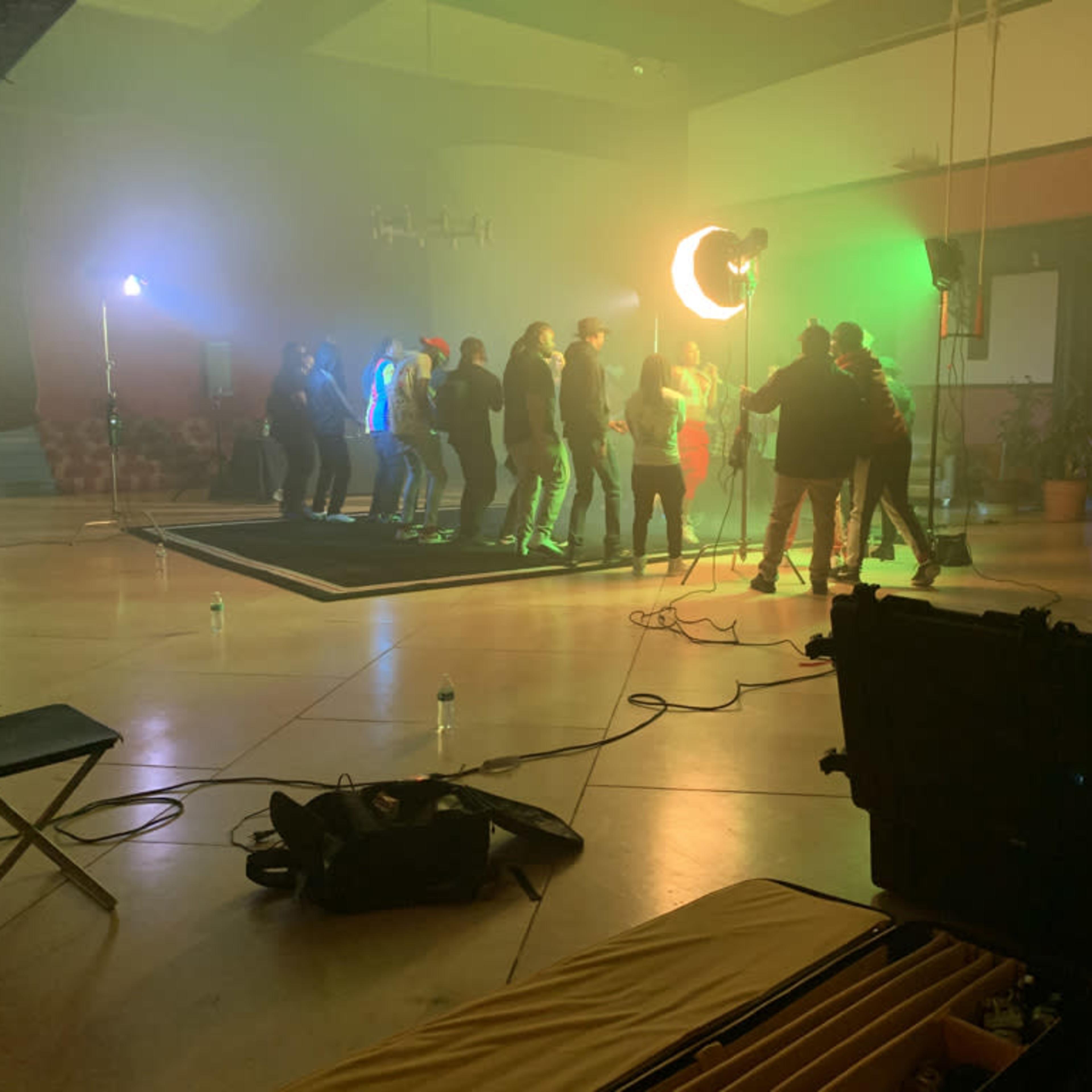 A group of people are gathered on a stage in a well-lit room with colorful lighting, while a film crew sets up equipment nearby.