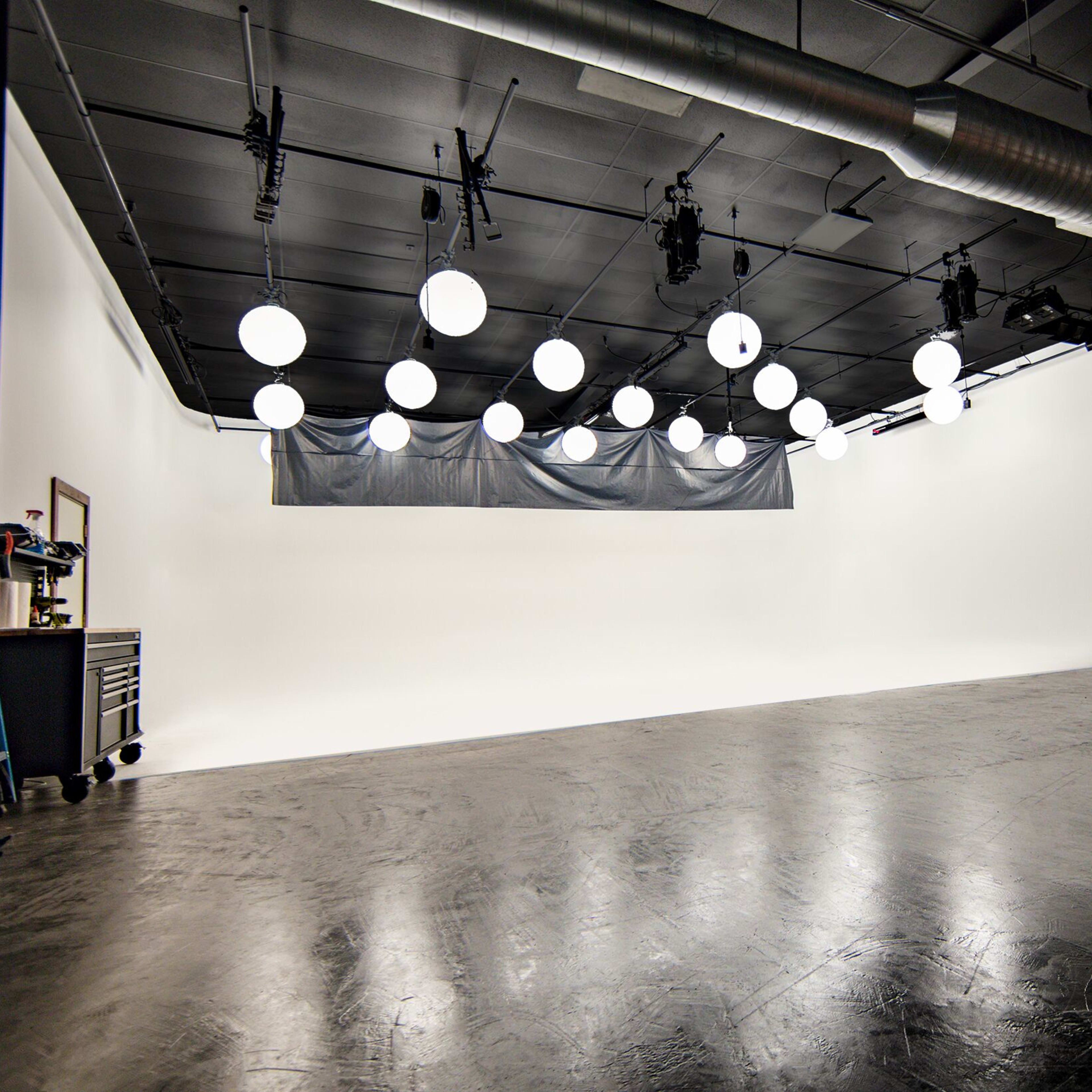 A photography studio with a blank white backdrop, dimly lit by multiple round lights hanging from the ceiling, and a brick wall on one side.