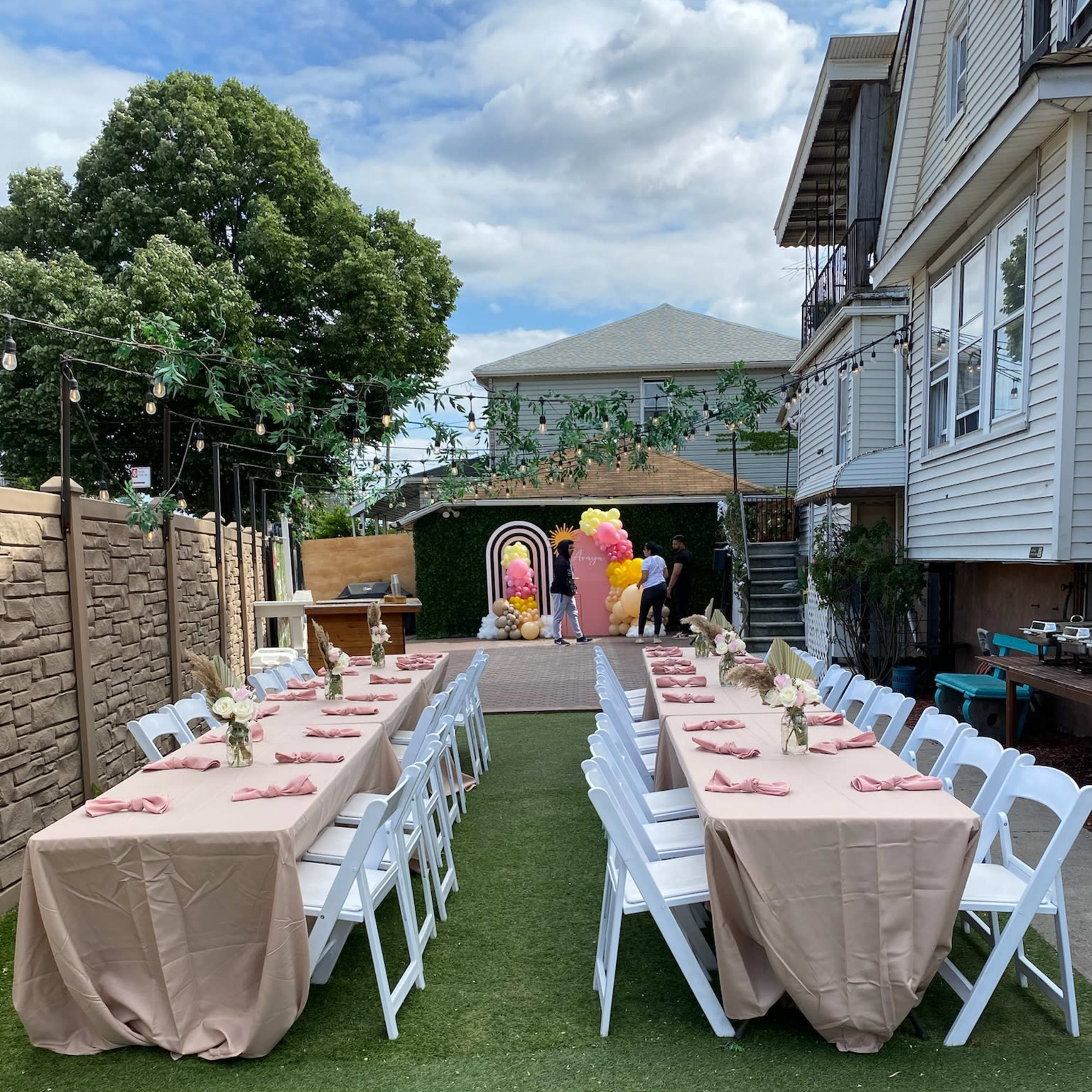A neatly arranged outdoor event space with long tables covered in beige tablecloths, set with pink napkins, surrounded by greenery and decorations.
