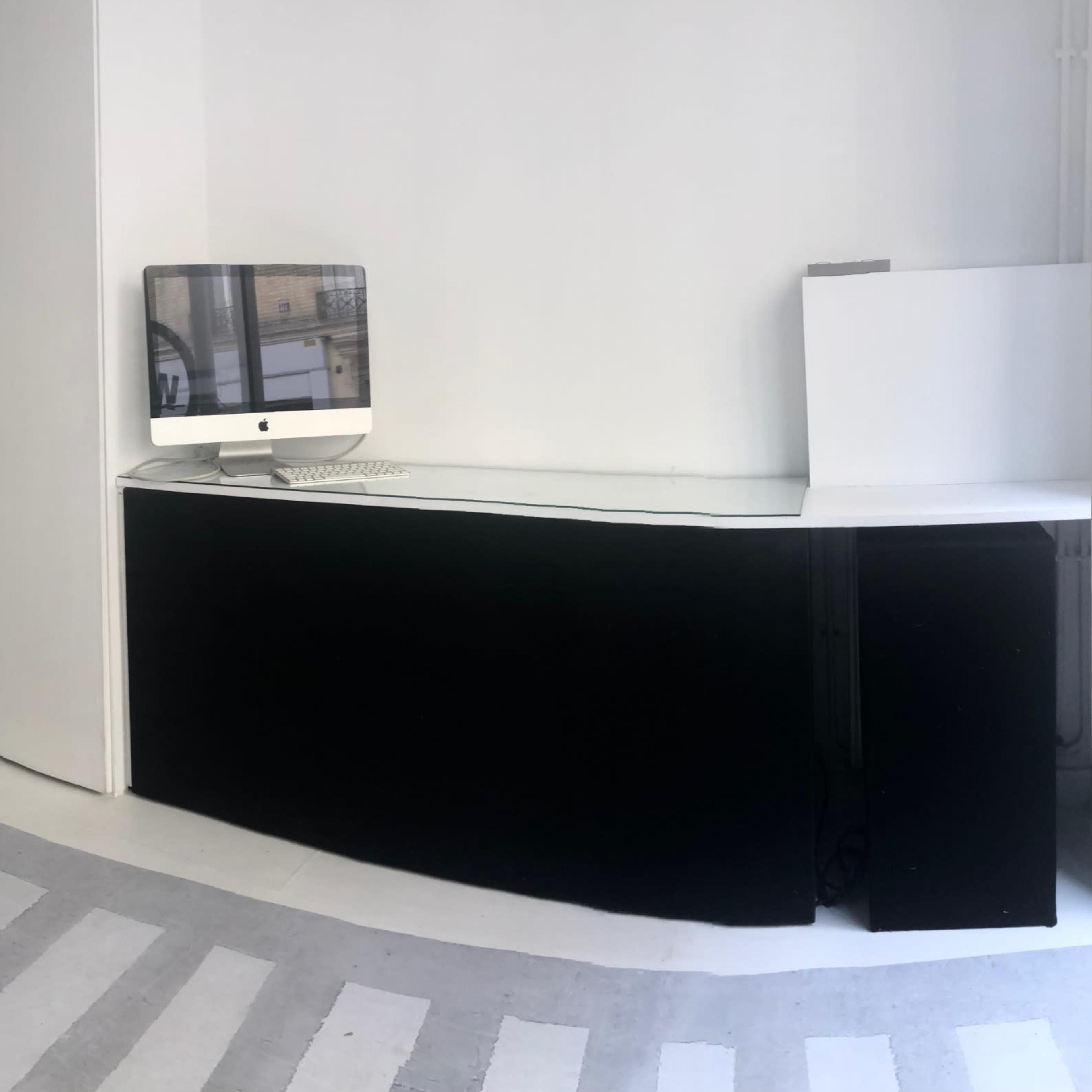 A minimalist, white-walled interior space featuring a reception area with a computer, a gray striped pedestrian walkway on the floor, and two black furniture pieces in the background.