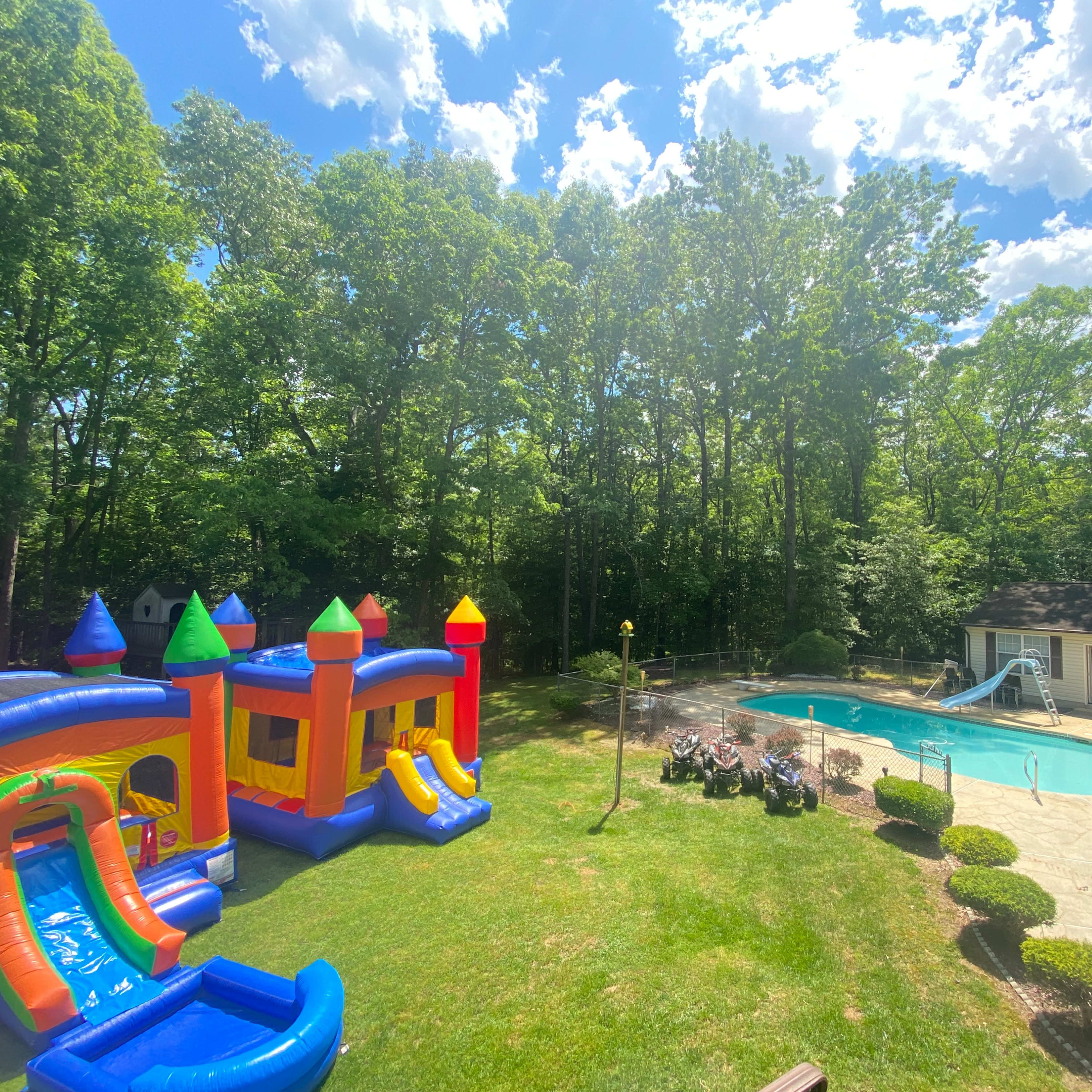 A backyard featuring a colorful inflatable bounce house and slide next to a swimming pool surrounded by trees.