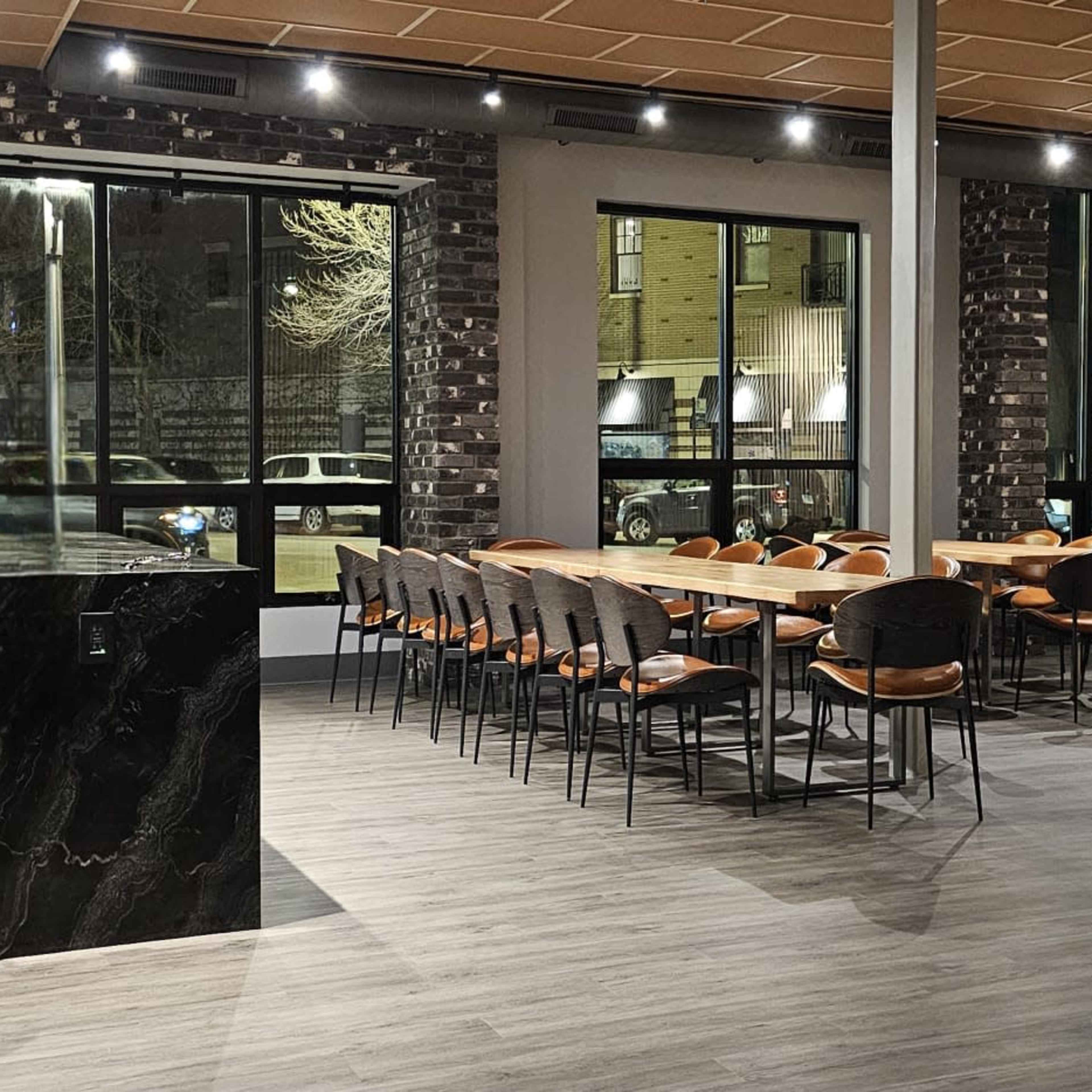 A spacious dining area with a long wooden table surrounded by chairs, large windows displaying a nighttime street view, and dark stone accents.
