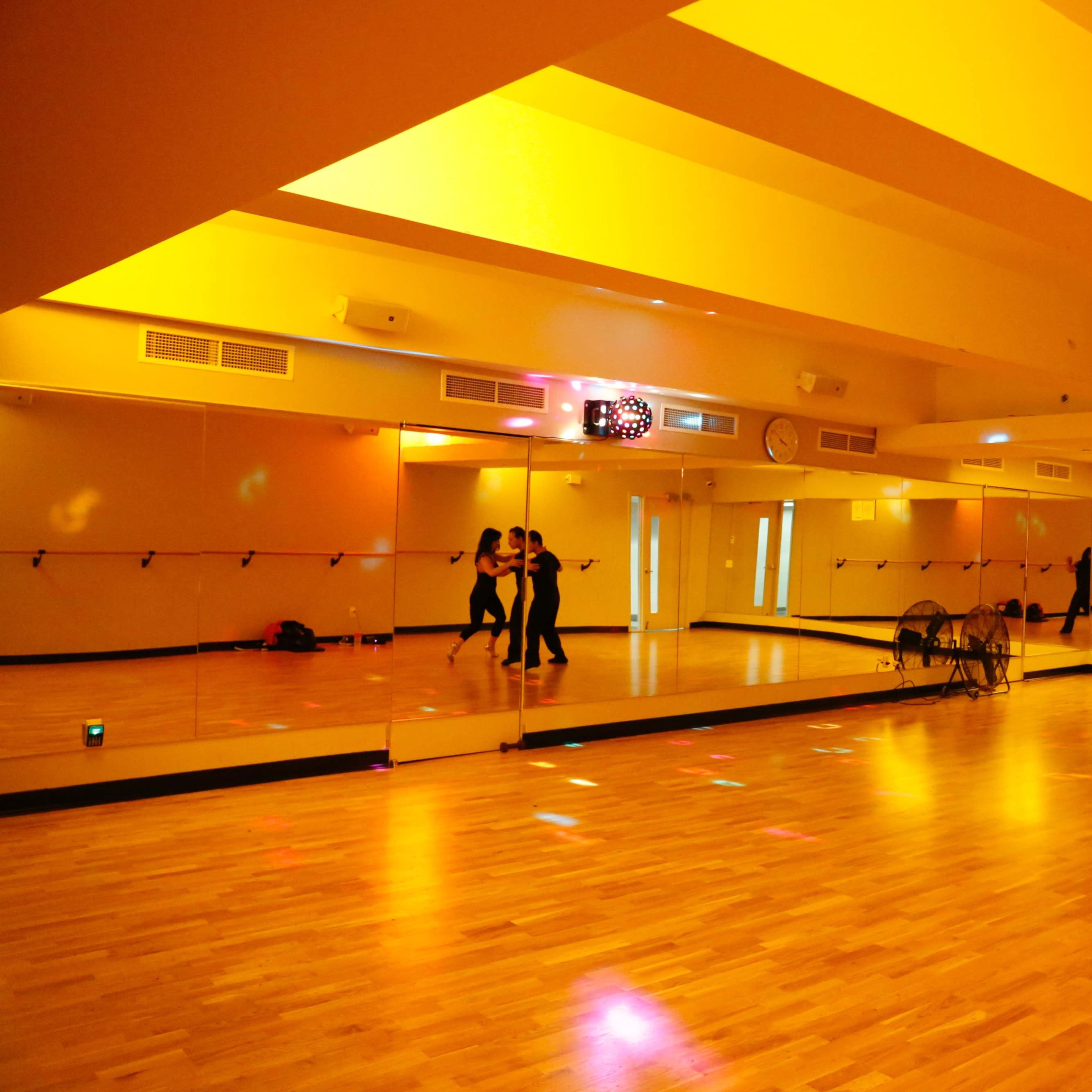 A dance studio with mirrored walls and warm yellow lighting features two dancers practicing in the center of the room.