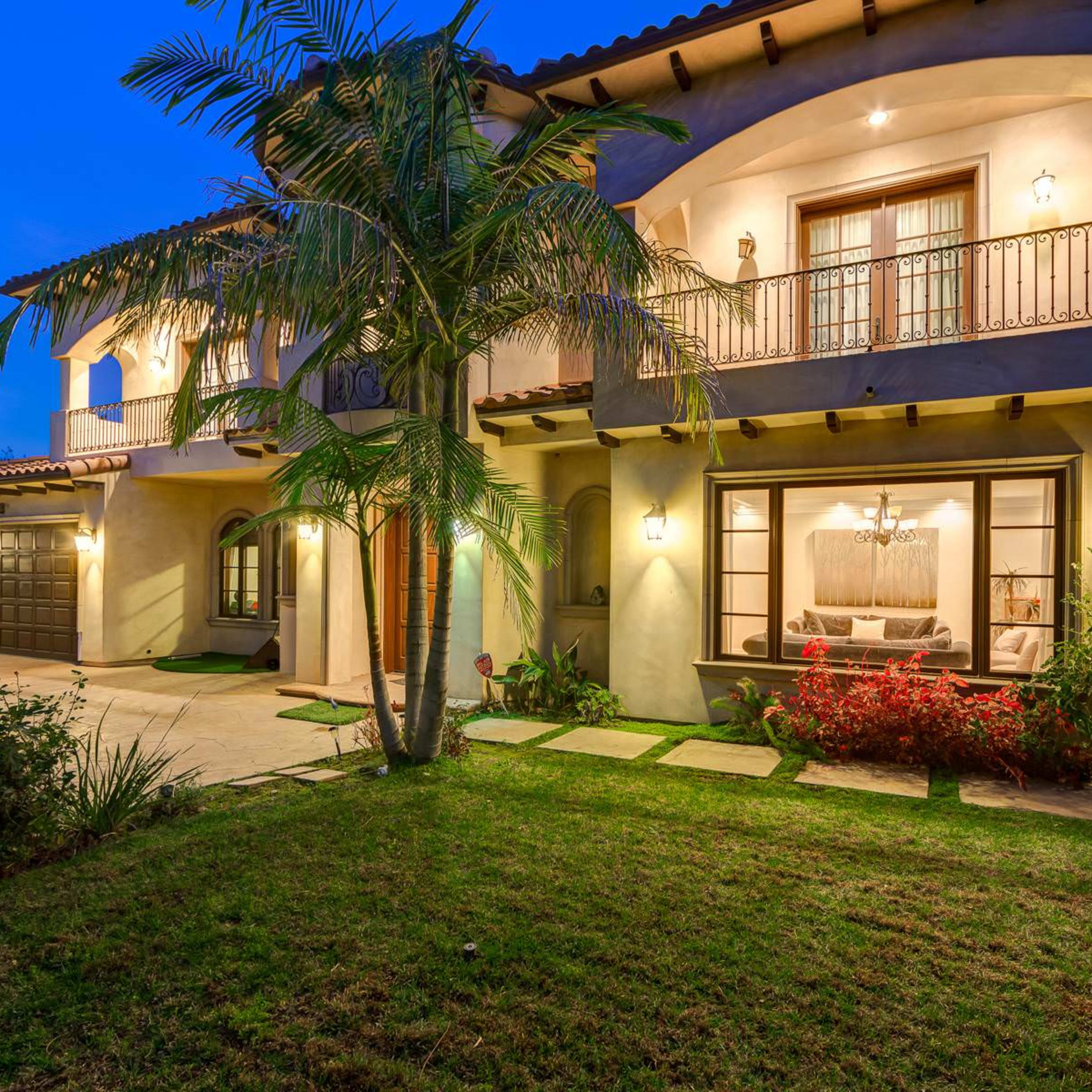 A two-story house with a balcony, surrounded by lush greenery, is illuminated at dusk.