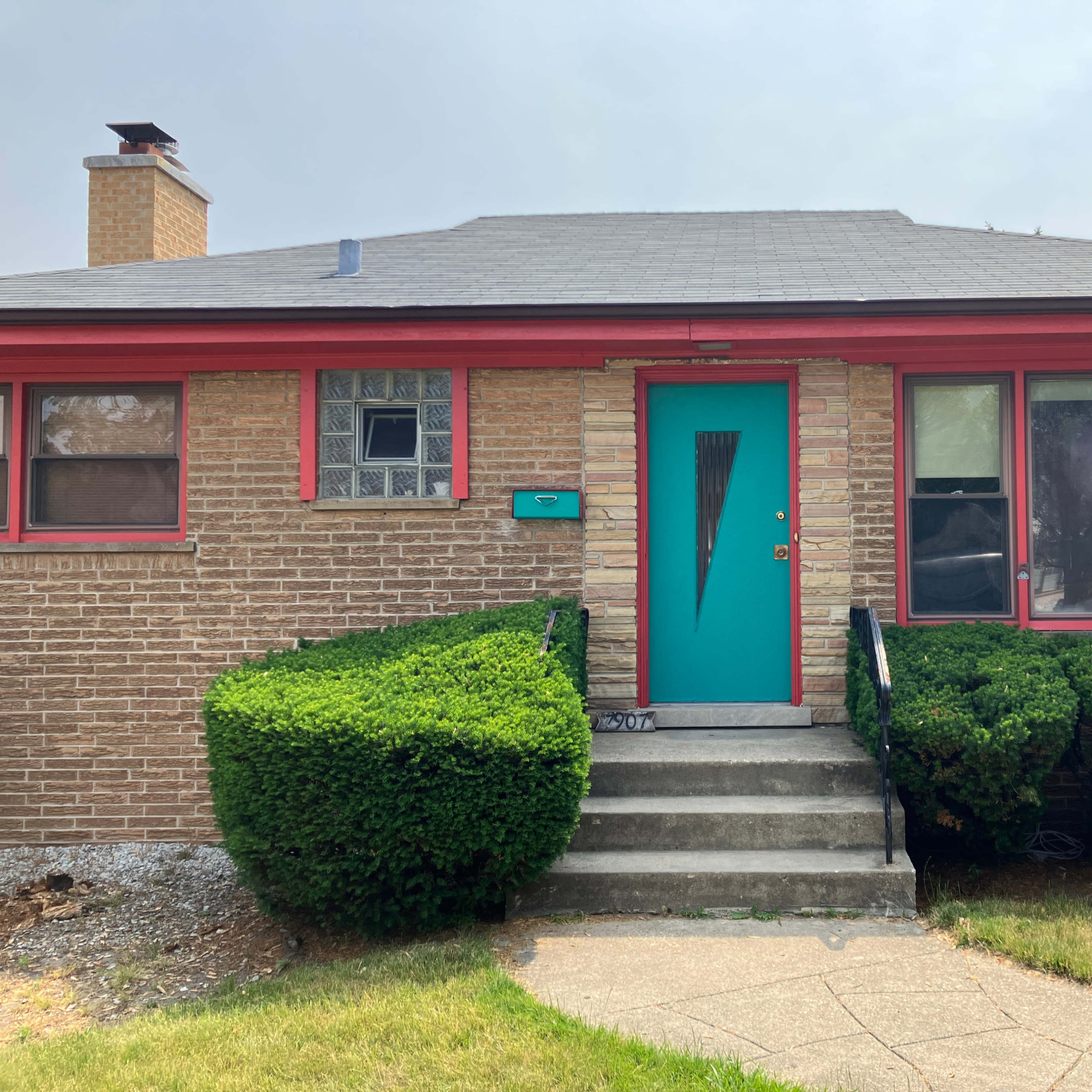 A bungalow-style house with a turquoise front door and vibrant red trim, surrounded by neatly trimmed bushes and a concrete pathway.