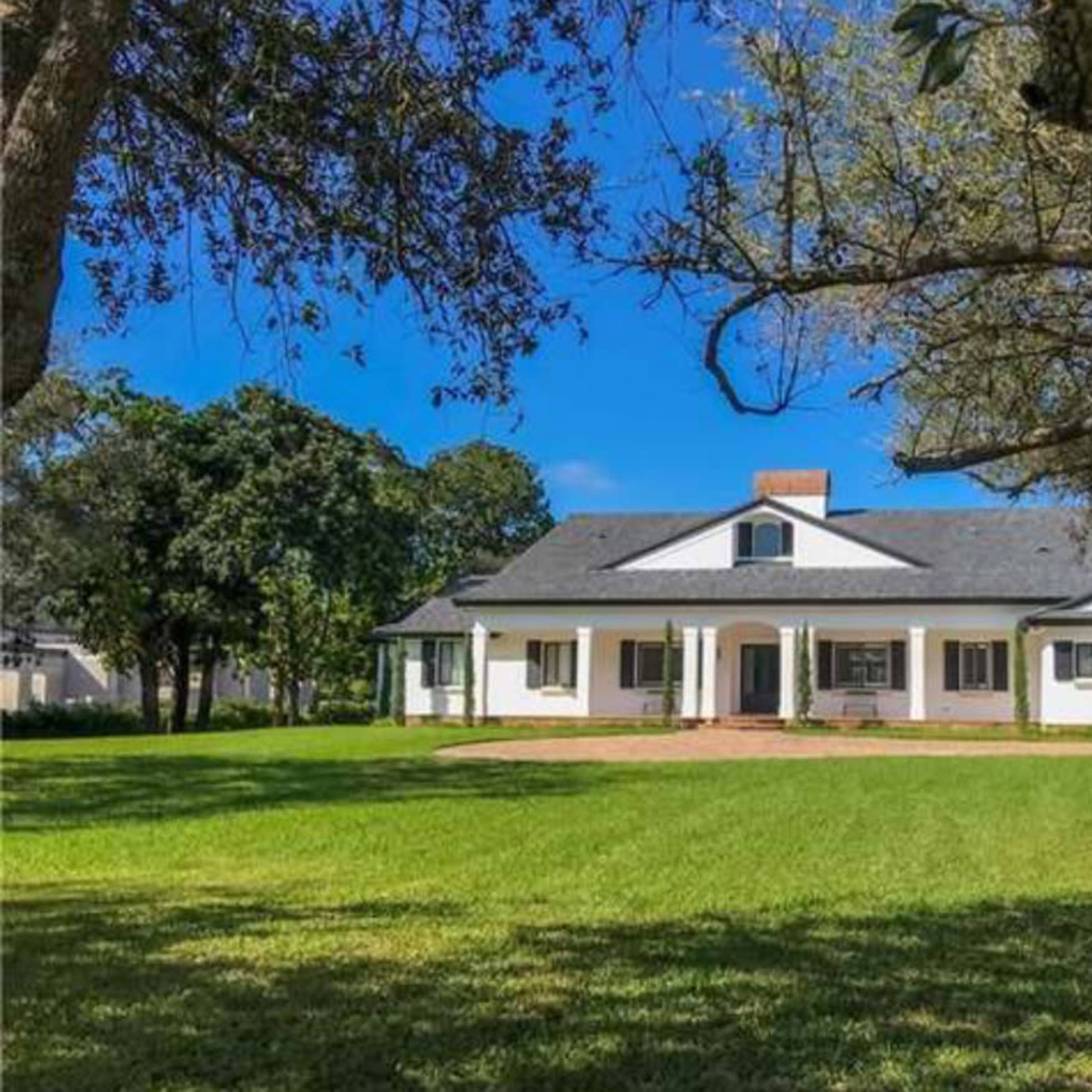 A single-story white house with a black roof and a central chimney sits on a green lawn surrounded by trees.