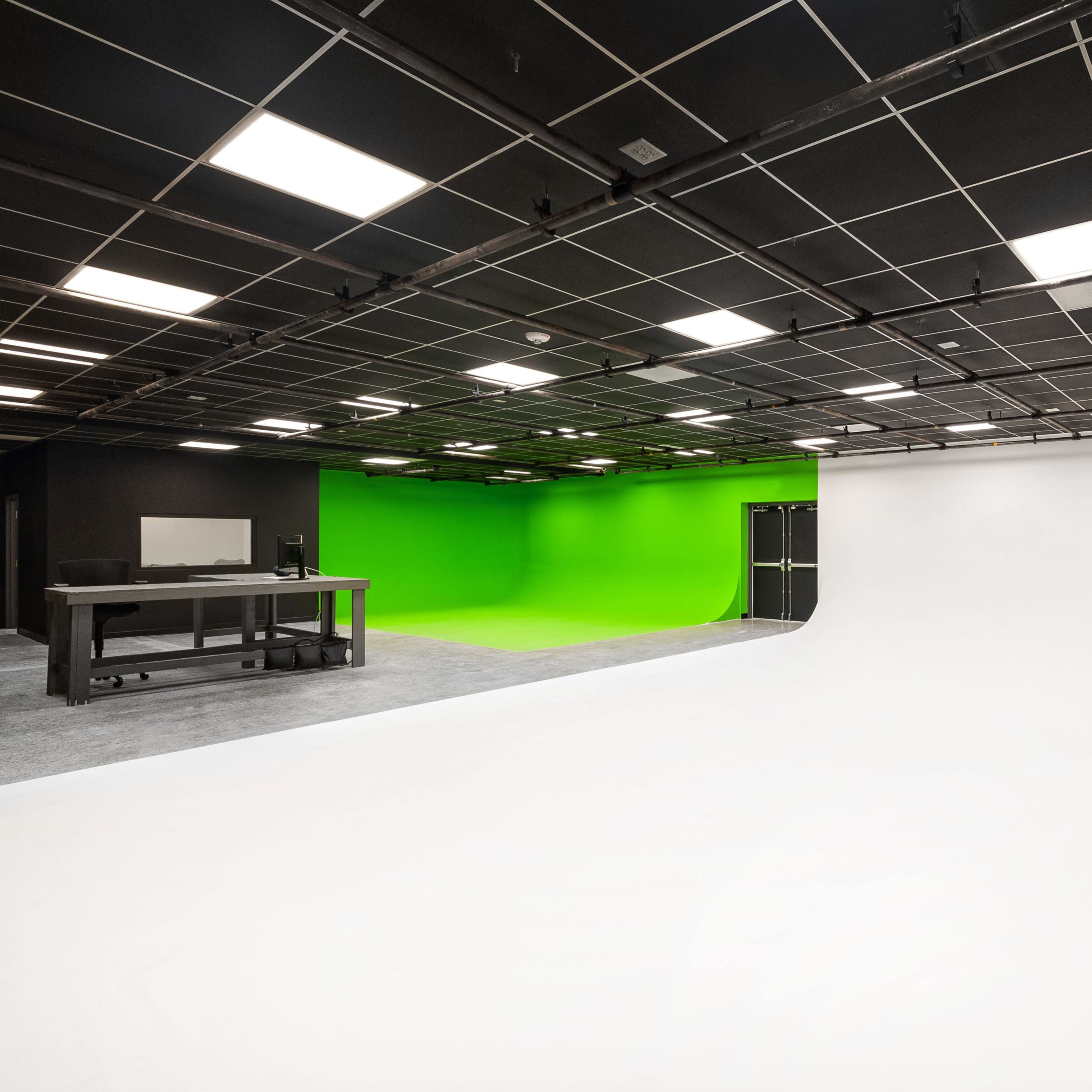 A spacious studio features a white background on one side and a green screen on the other, with a black ceiling and overhead lights.