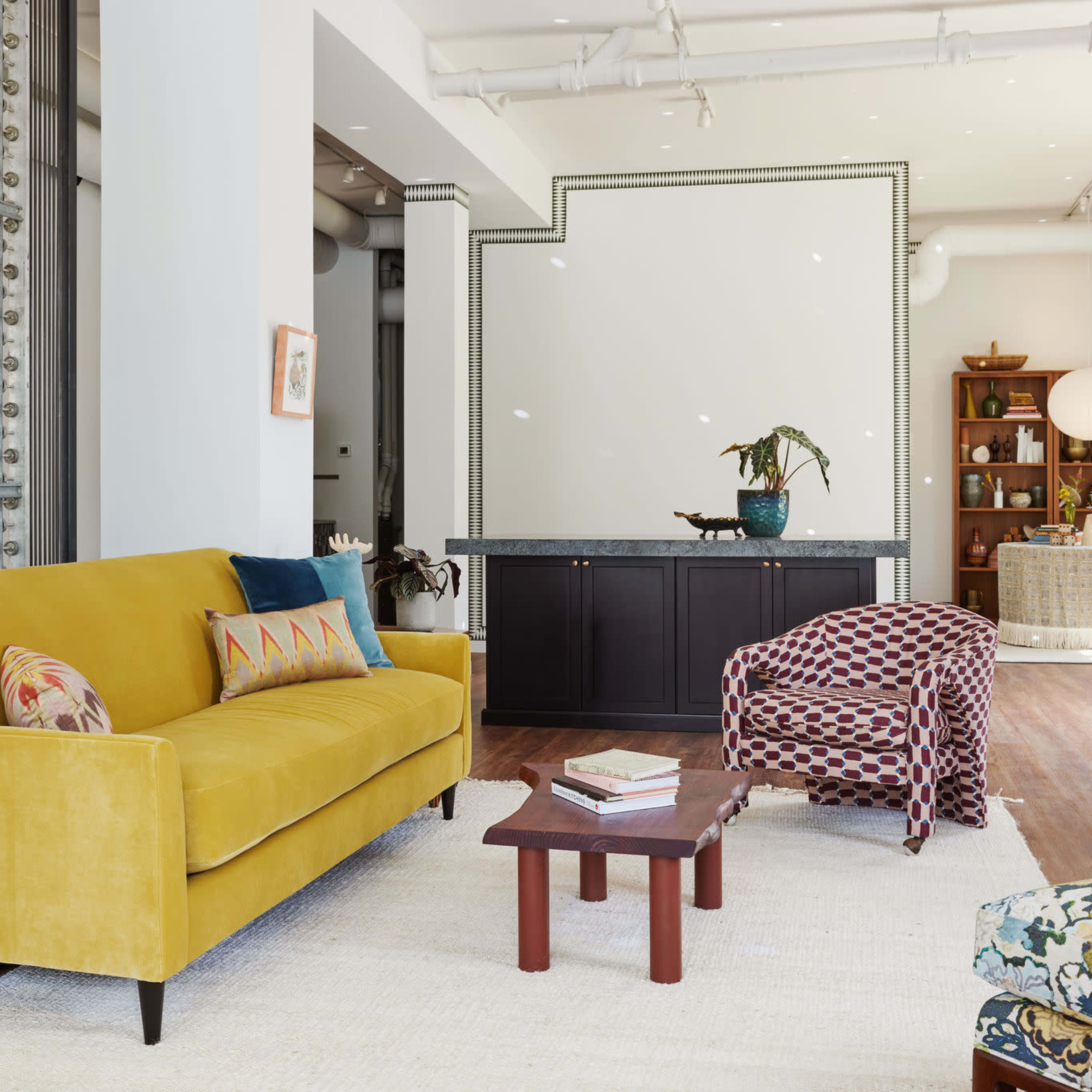A bright living room features a yellow sofa, patterned armchair, wooden coffee table, and a large white rug.