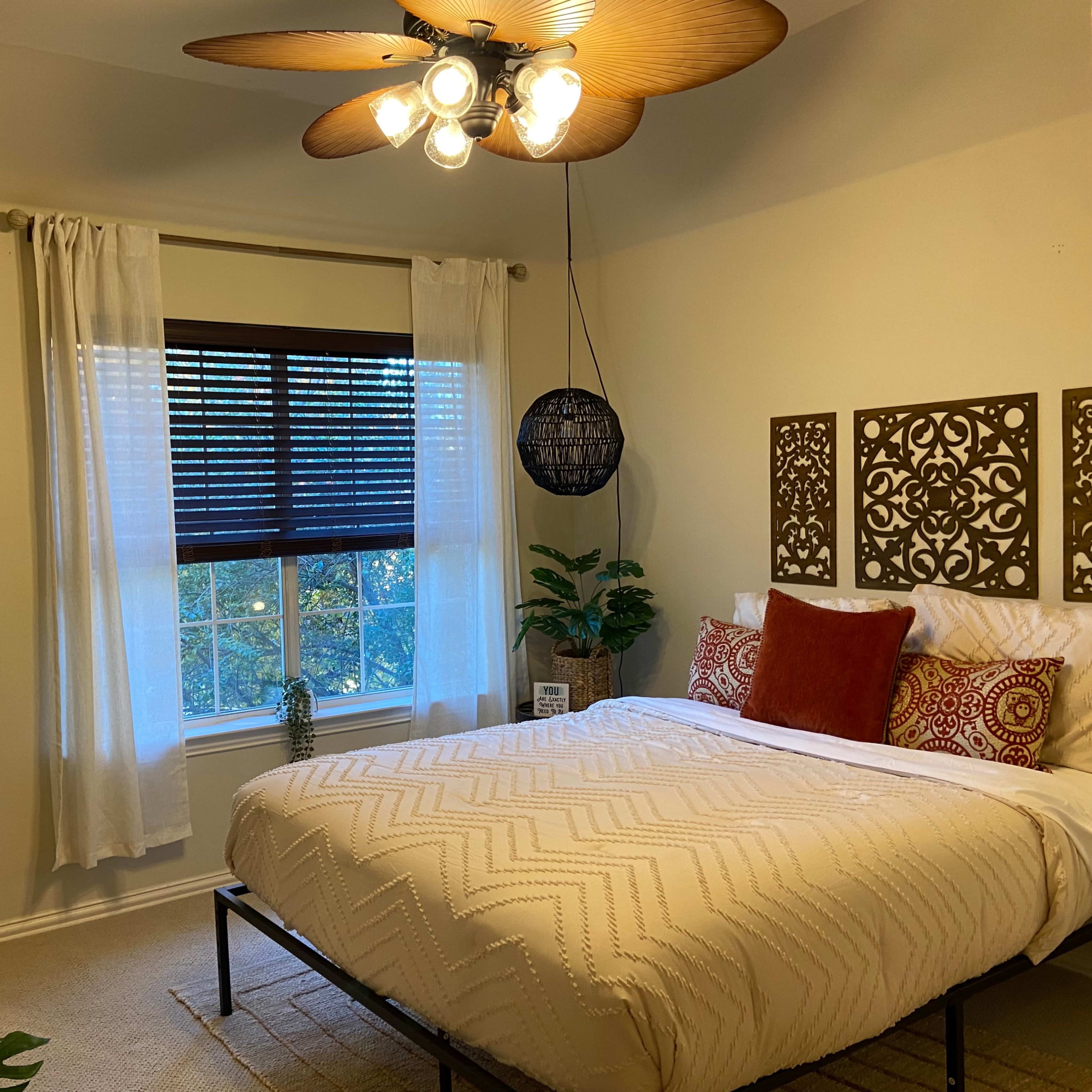 A well-decorated bedroom features a bed with patterned bedding, a large mirror, and a window with venetian blinds, complemented by plants and a ceiling fan.