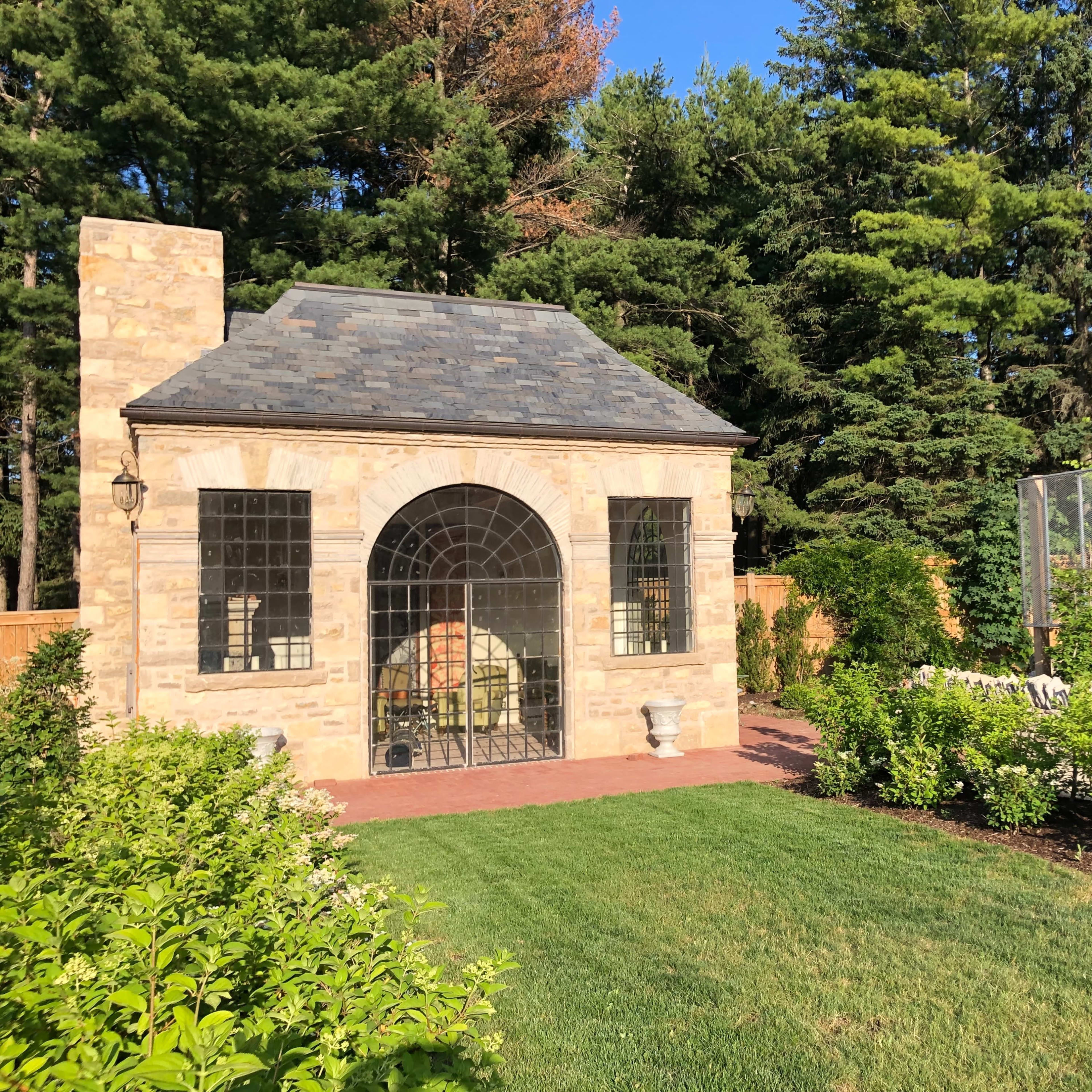 A stone garden structure with a large arched window surrounded by manicured greenery and trees.