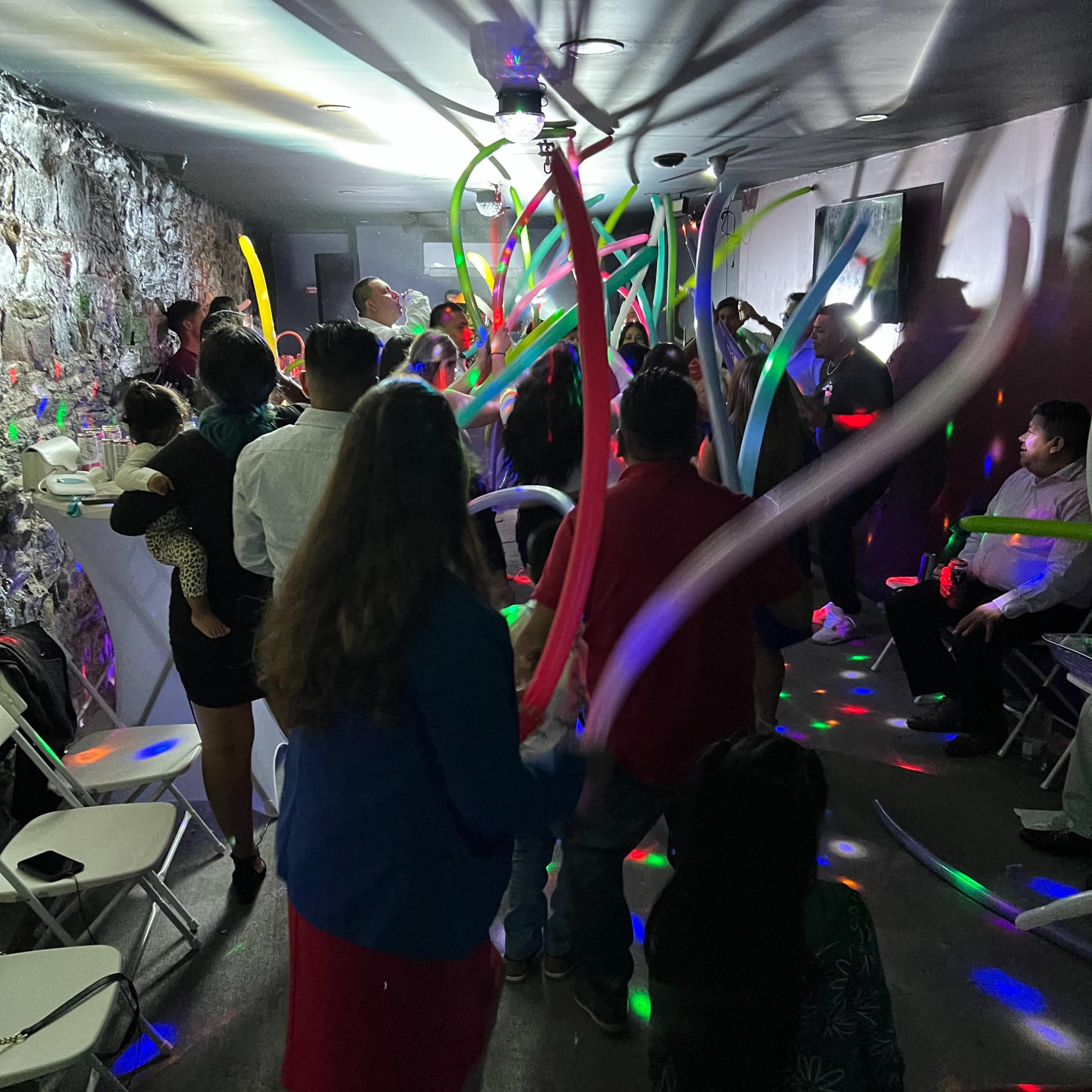 A group of people is dancing in a dimly lit room with colorful lights and long balloons swirling above them.