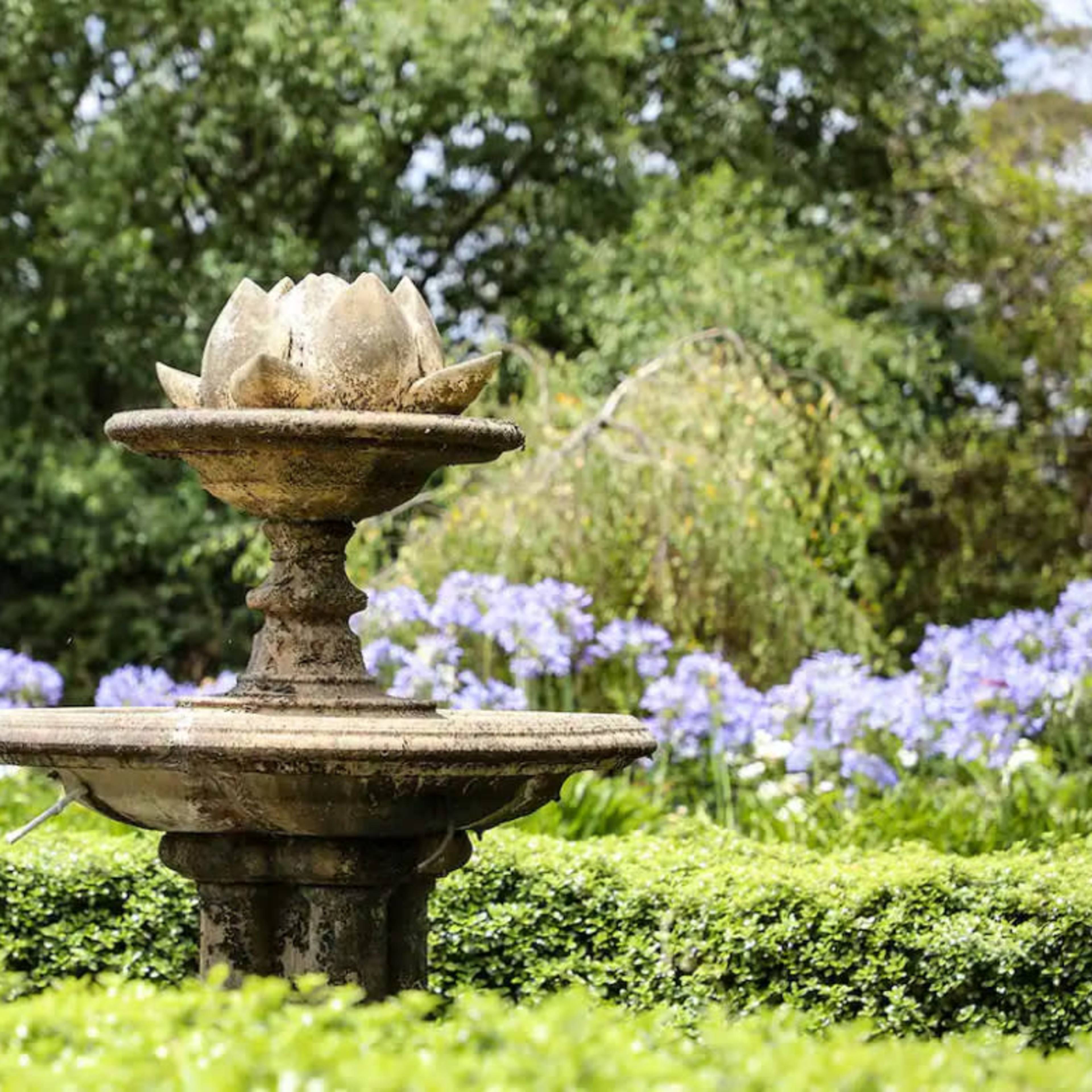 A stone fountain shaped like a lotus flower is surrounded by greenery and blooming purple flowers.
