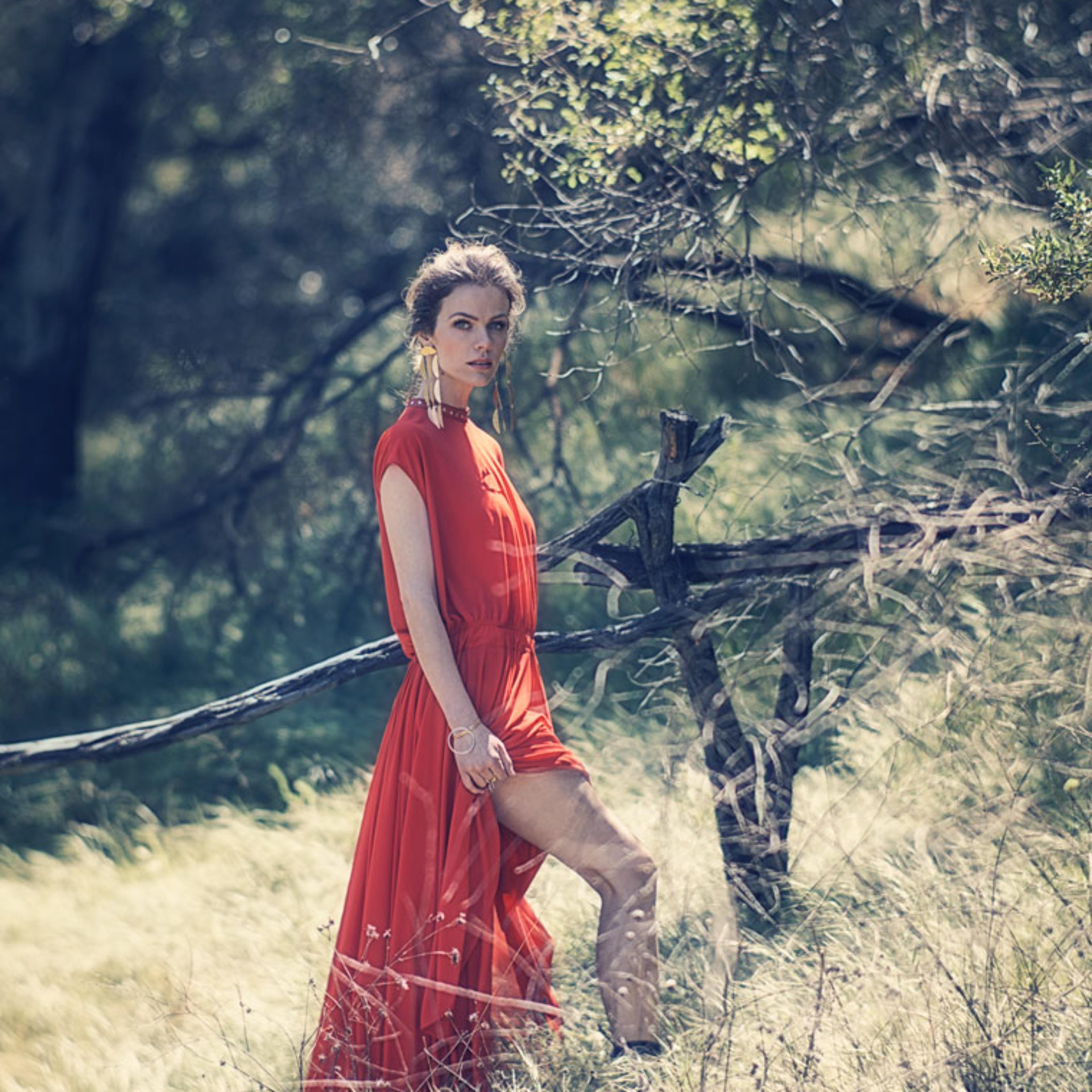 A woman in a red dress stands among tall grass and trees in a natural setting.