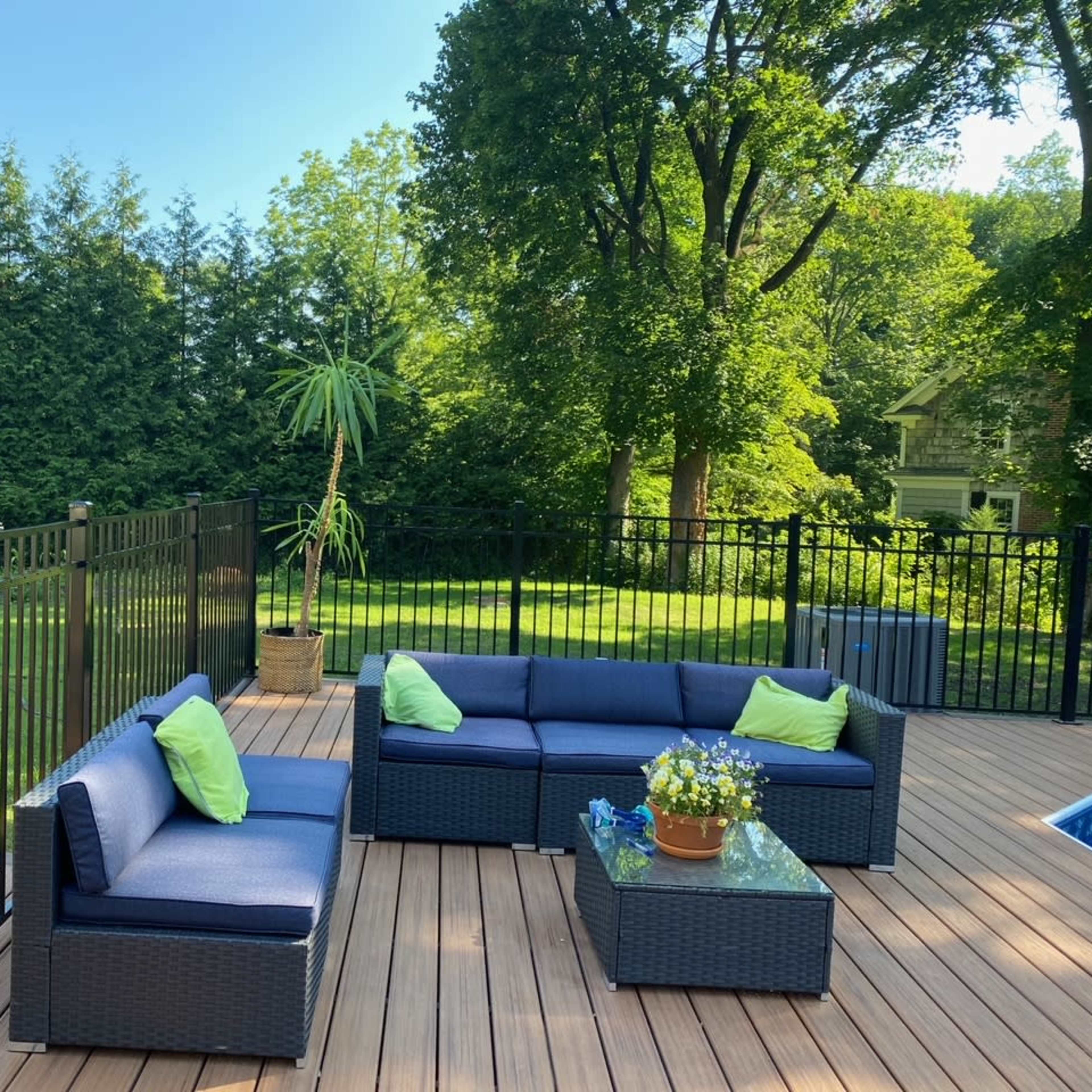 A patio area features dark wicker furniture with green cushions, surrounded by a wooden deck and green trees in the background.
