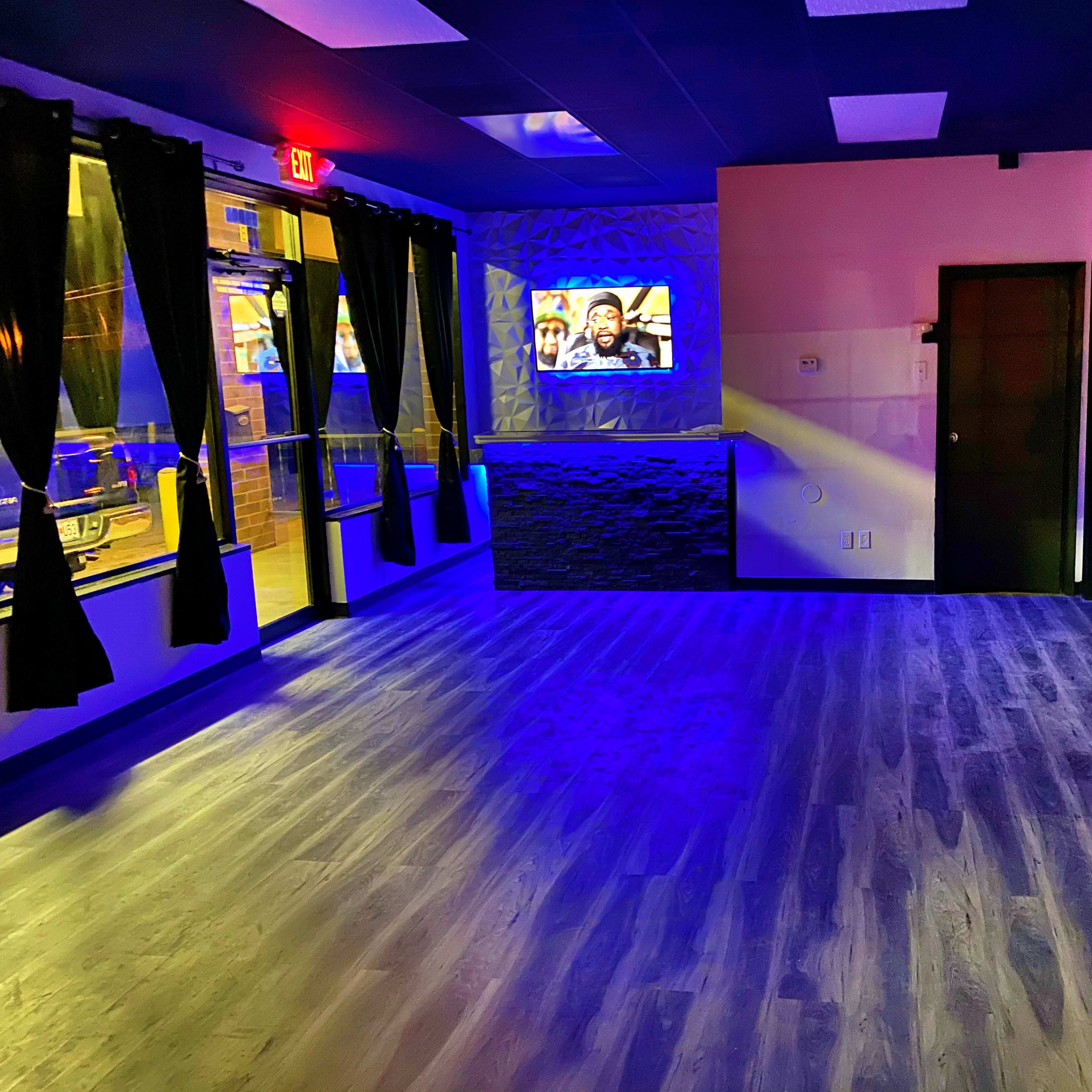 An empty room with blue lighting, featuring a textured wall, a television, large windows with dark curtains, and a mirror.