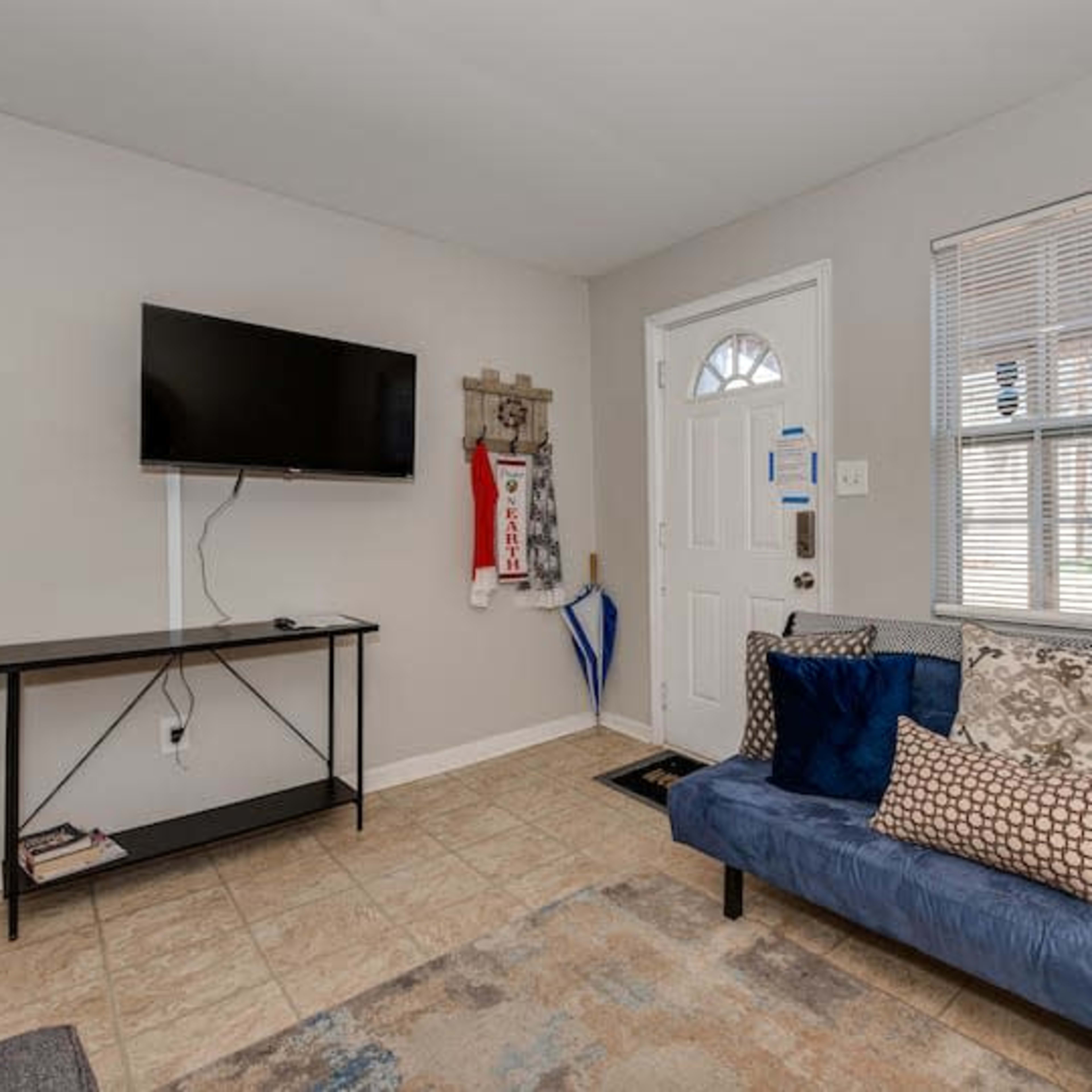 The room features a blue couch with decorative pillows, a wall-mounted television, and a door leading outside.