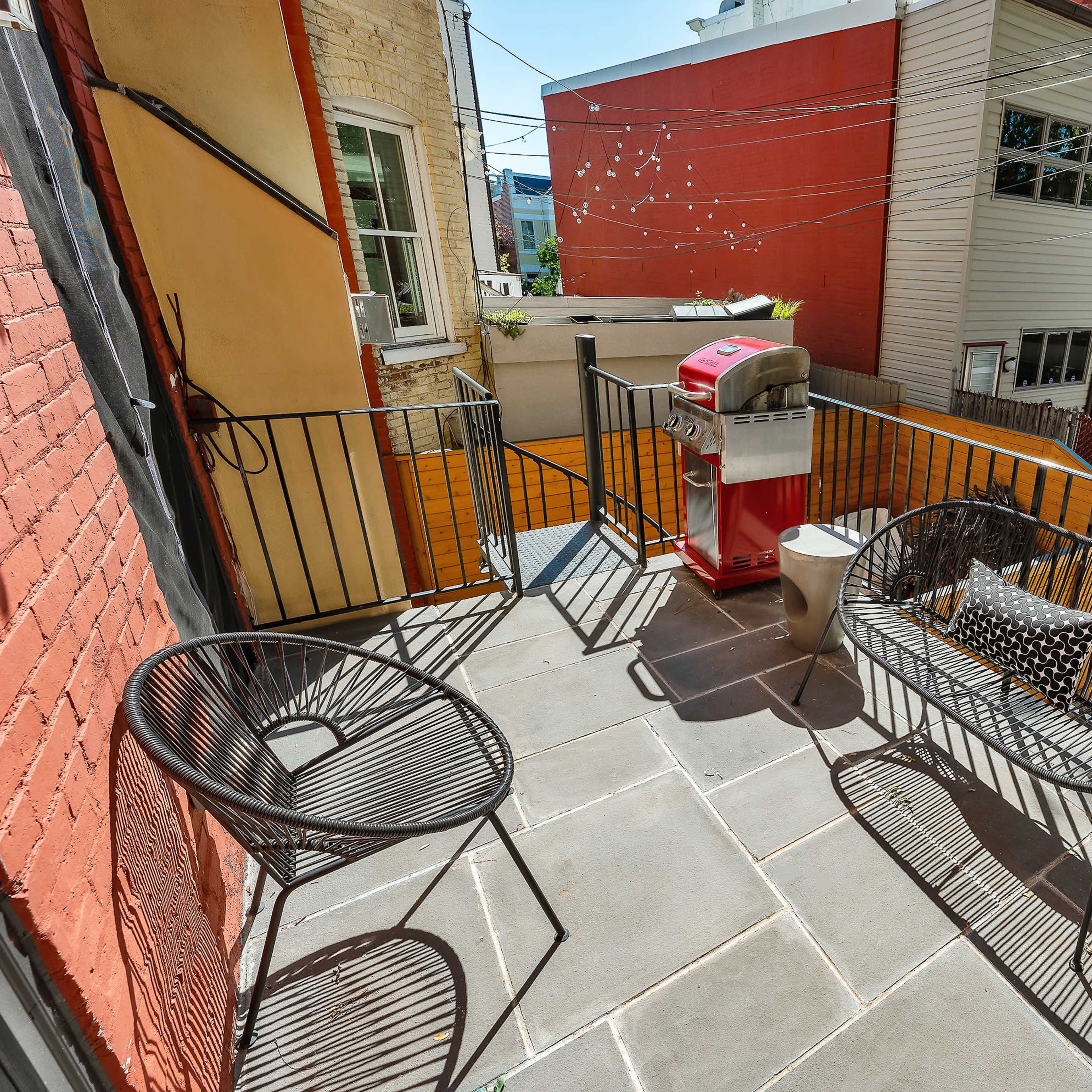 A balcony features two wire chairs, a small table, and a red grill, with views of surrounding buildings and a courtyard.
