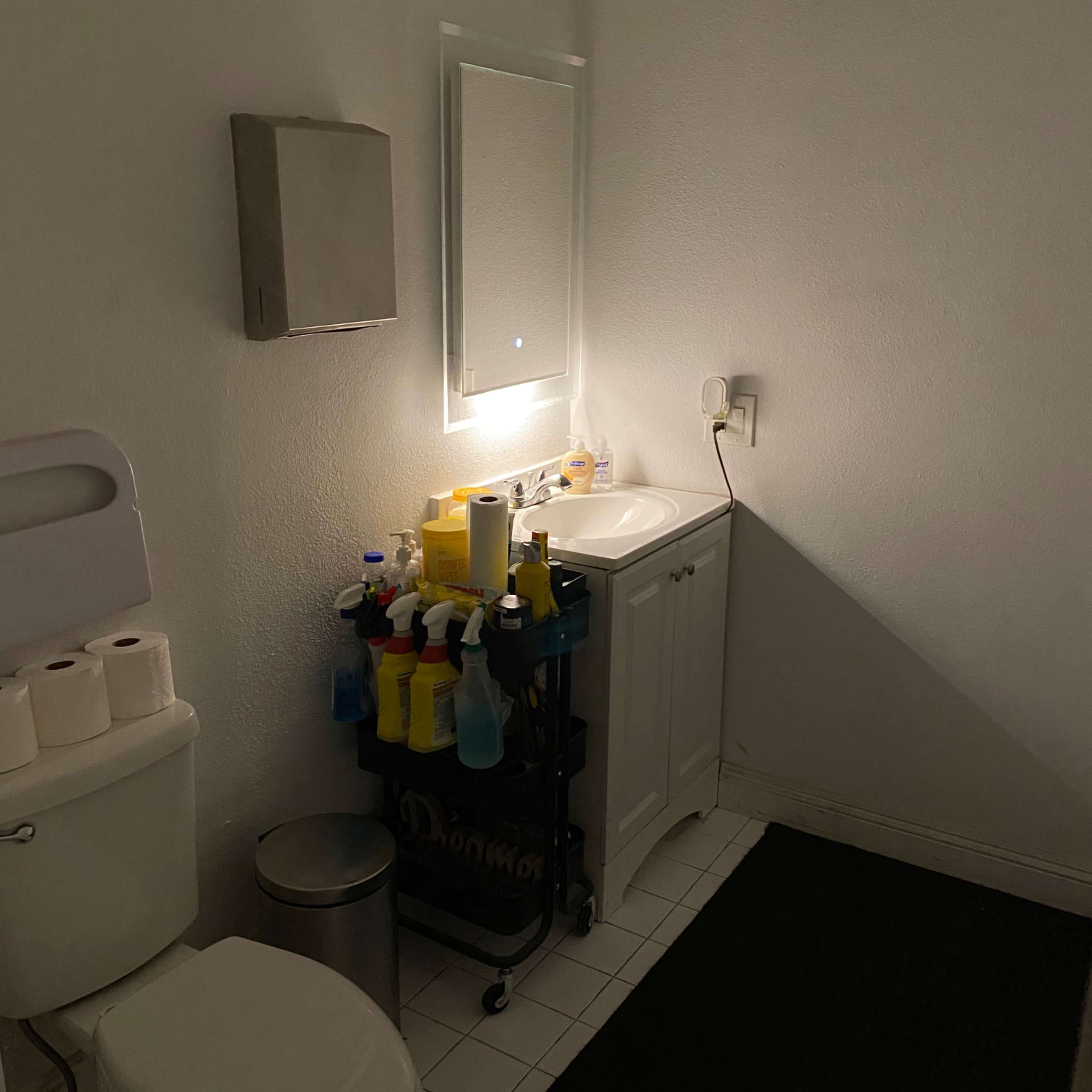 A dimly lit bathroom features a toilet, a small sink with a mirror, and a cart filled with cleaning supplies.
