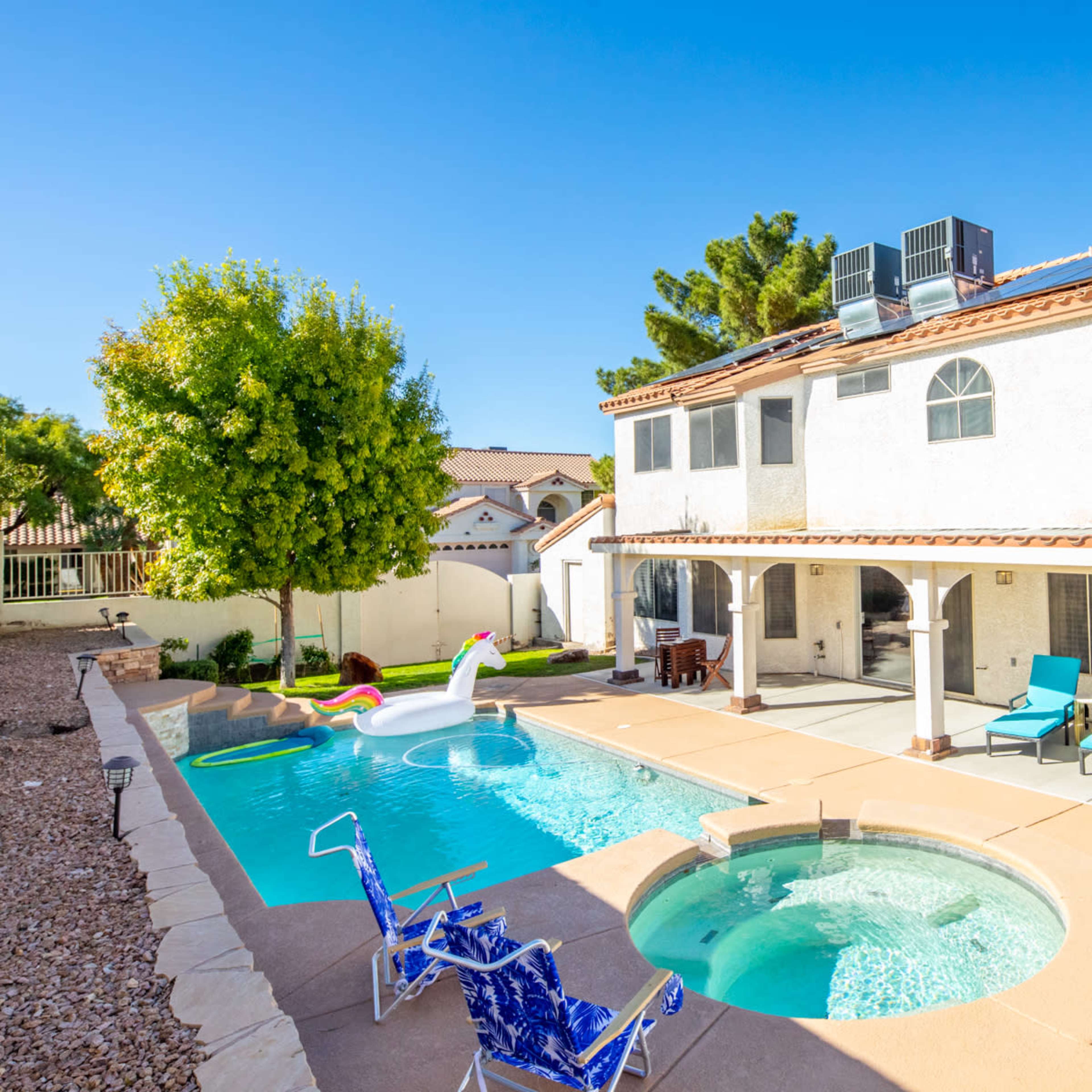 A two-story house with a swimming pool, spa, and lounge chairs in the backyard, surrounded by palm trees and residential homes.