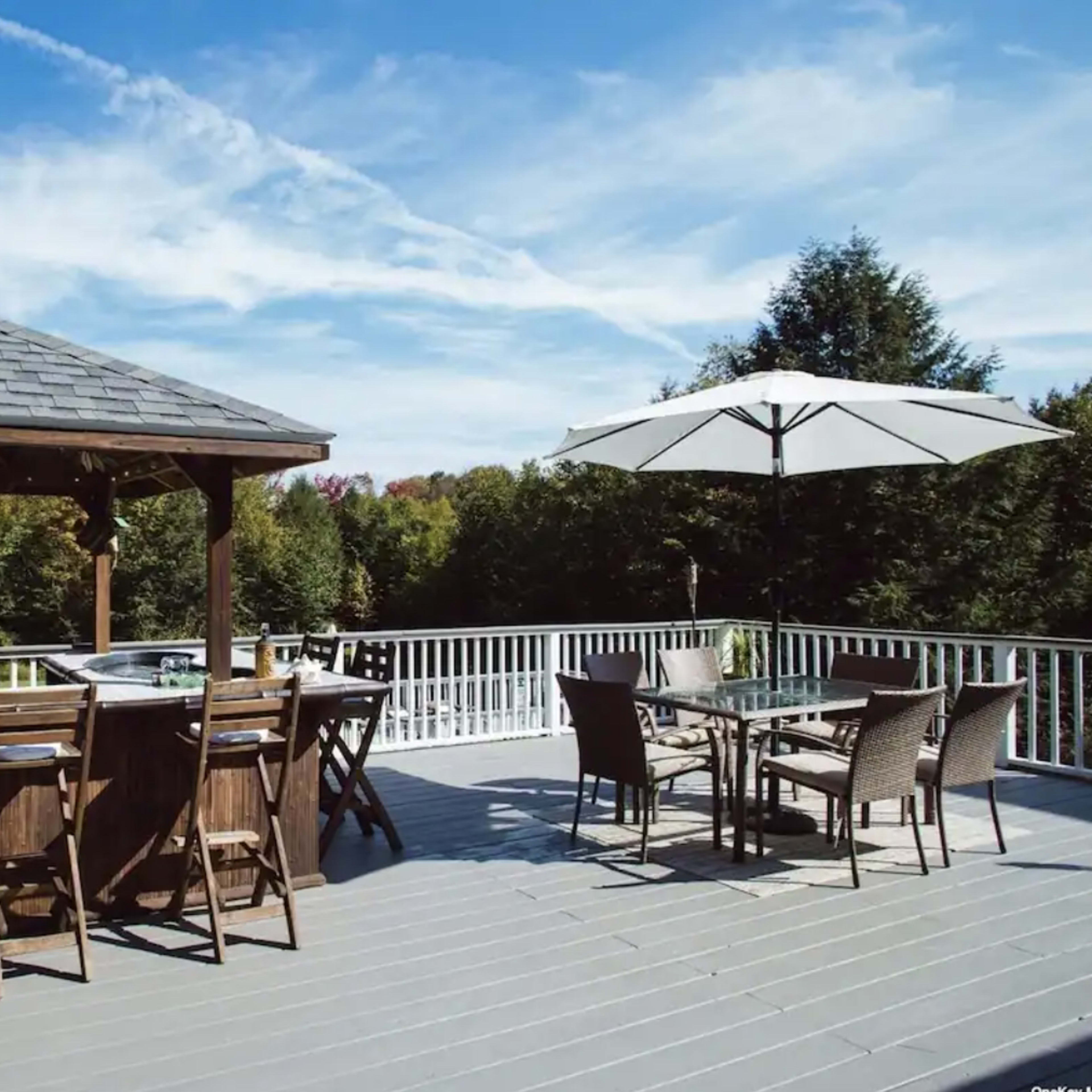 A wooden deck features a covered bar area with stools, a dining table with chairs, and a large umbrella, surrounded by trees under a blue sky.