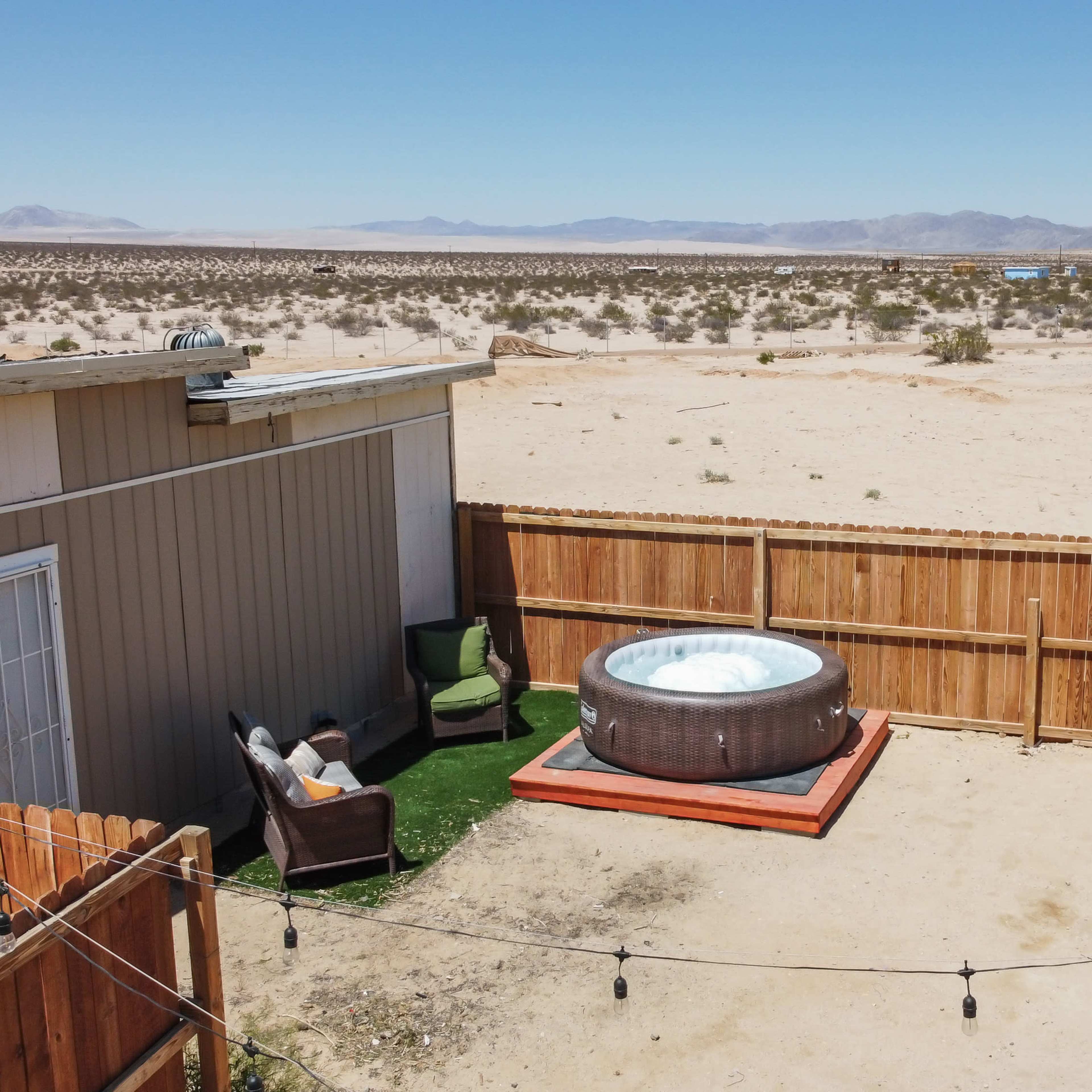 A fenced outdoor area in a desert landscape, featuring a hot tub on a wooden deck and a couple of chairs nearby.