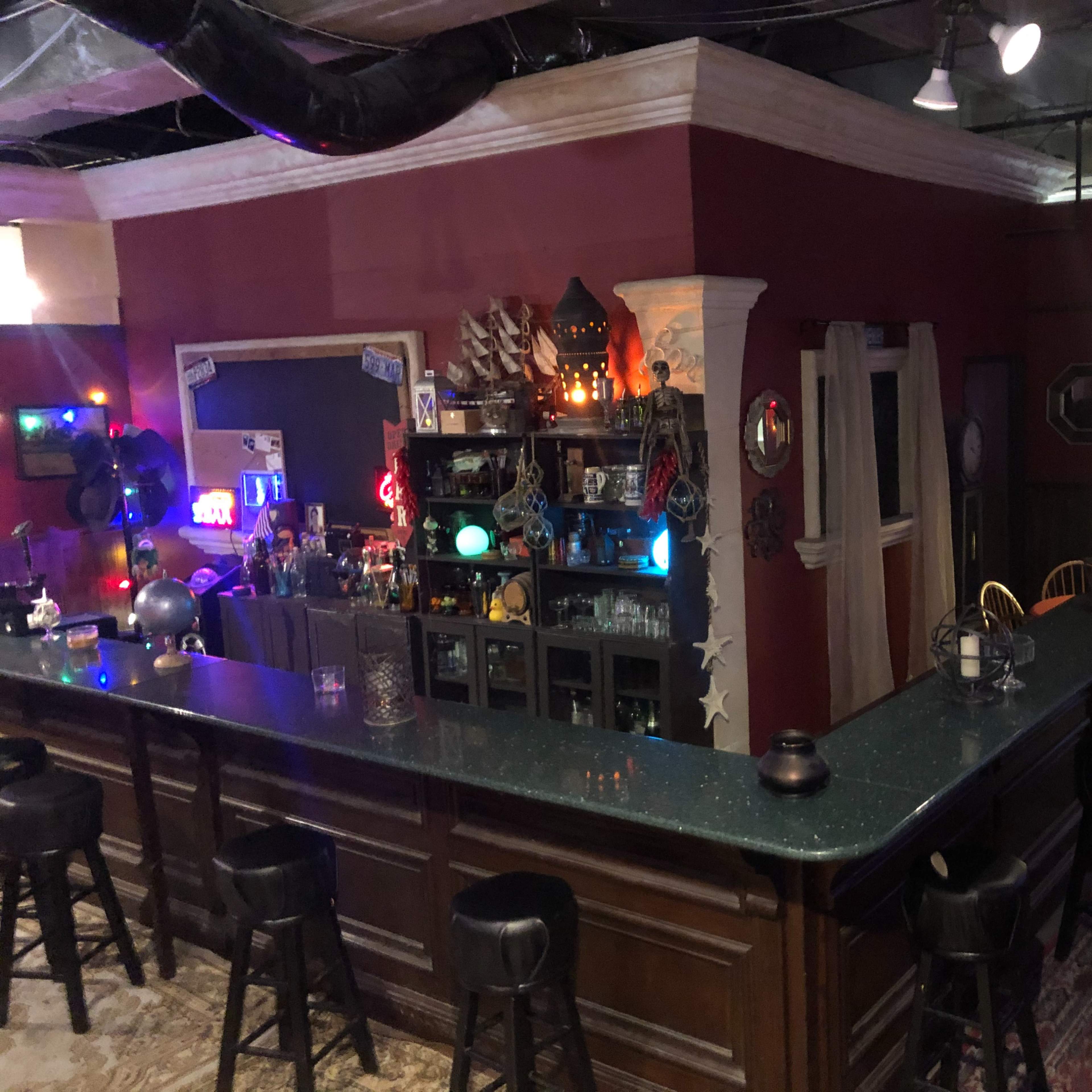 A dimly lit bar area with a large countertop, shelves filled with decorative items, and colorful lights along the walls.