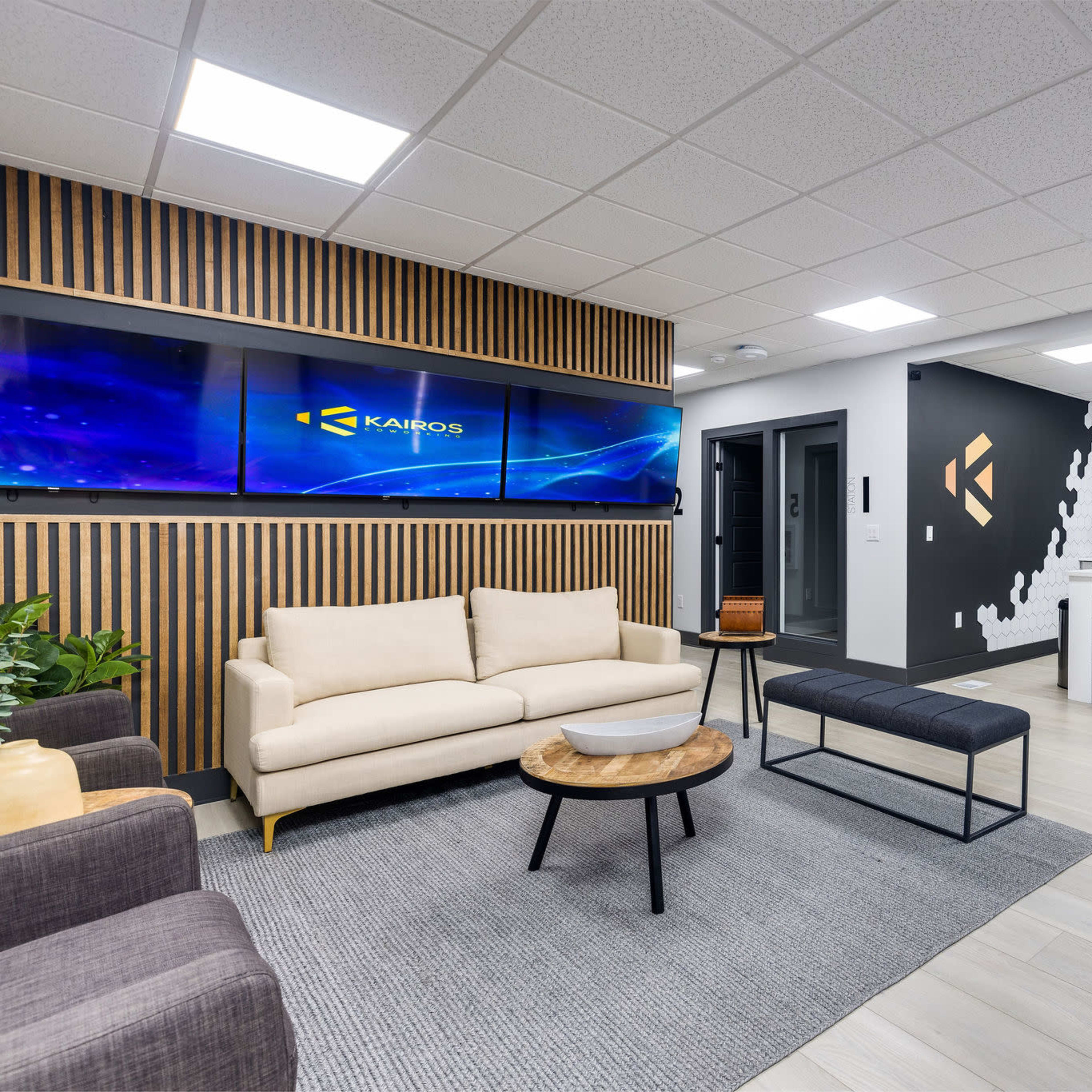 A modern office space with a seating area, a kitchen bar, and wall-mounted screens displaying the company logo against a backdrop of wooden slats and geometric designs.