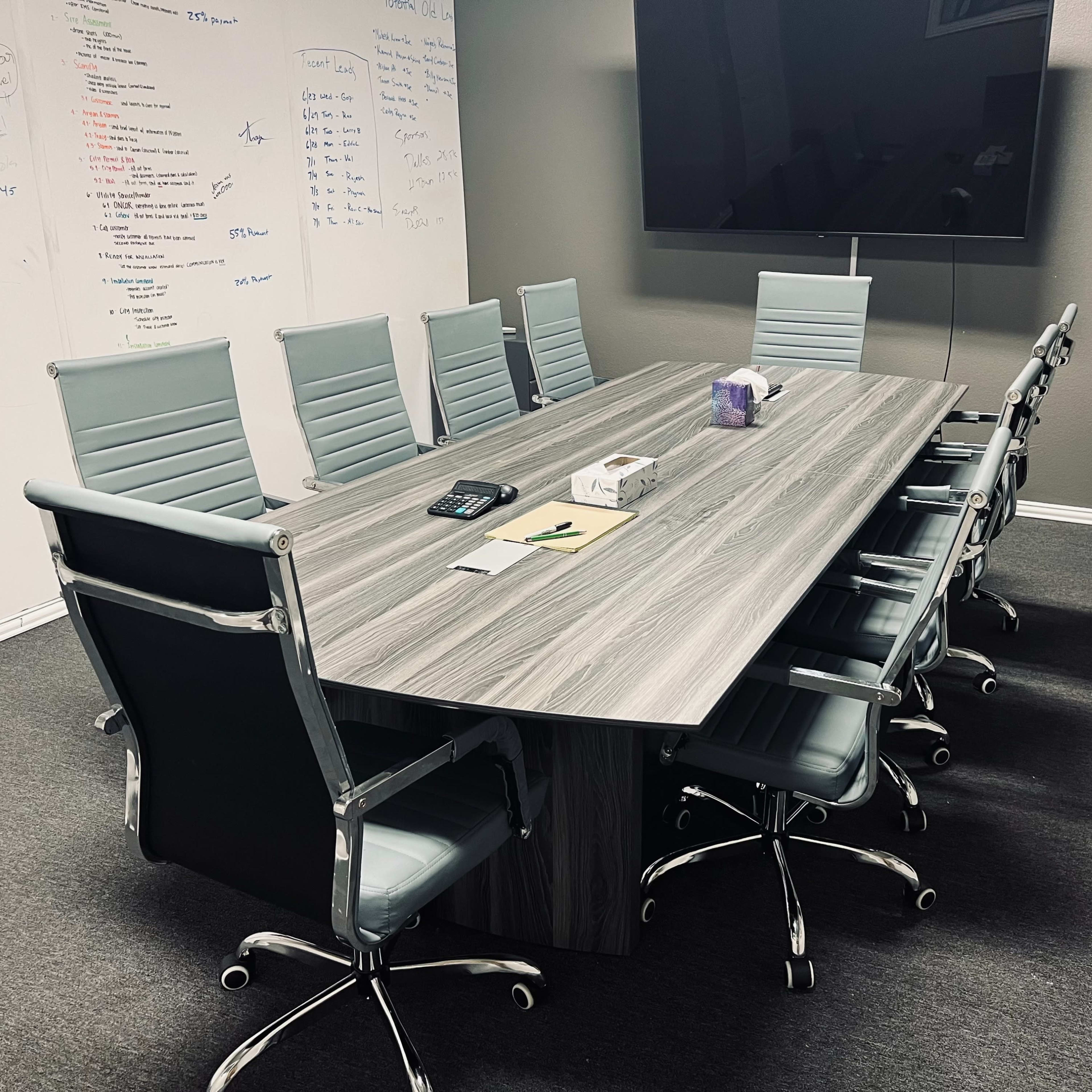 A modern conference room features a large table surrounded by multiple ergonomic chairs, with a wall-mounted screen and whiteboard covered in notes.