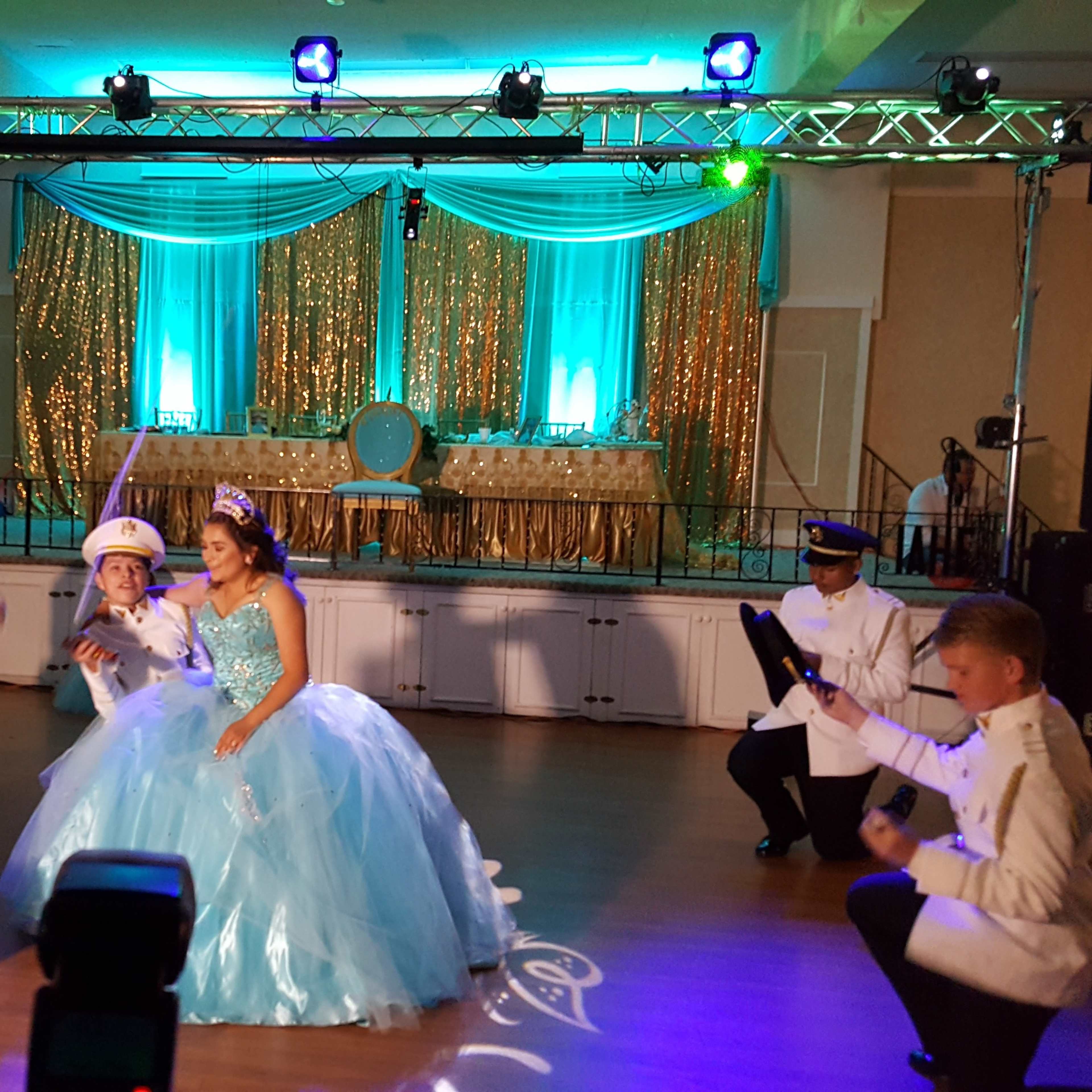 A group of young adults in formal attire perform a dance for a girl in a blue ball gown at a decorated event venue.