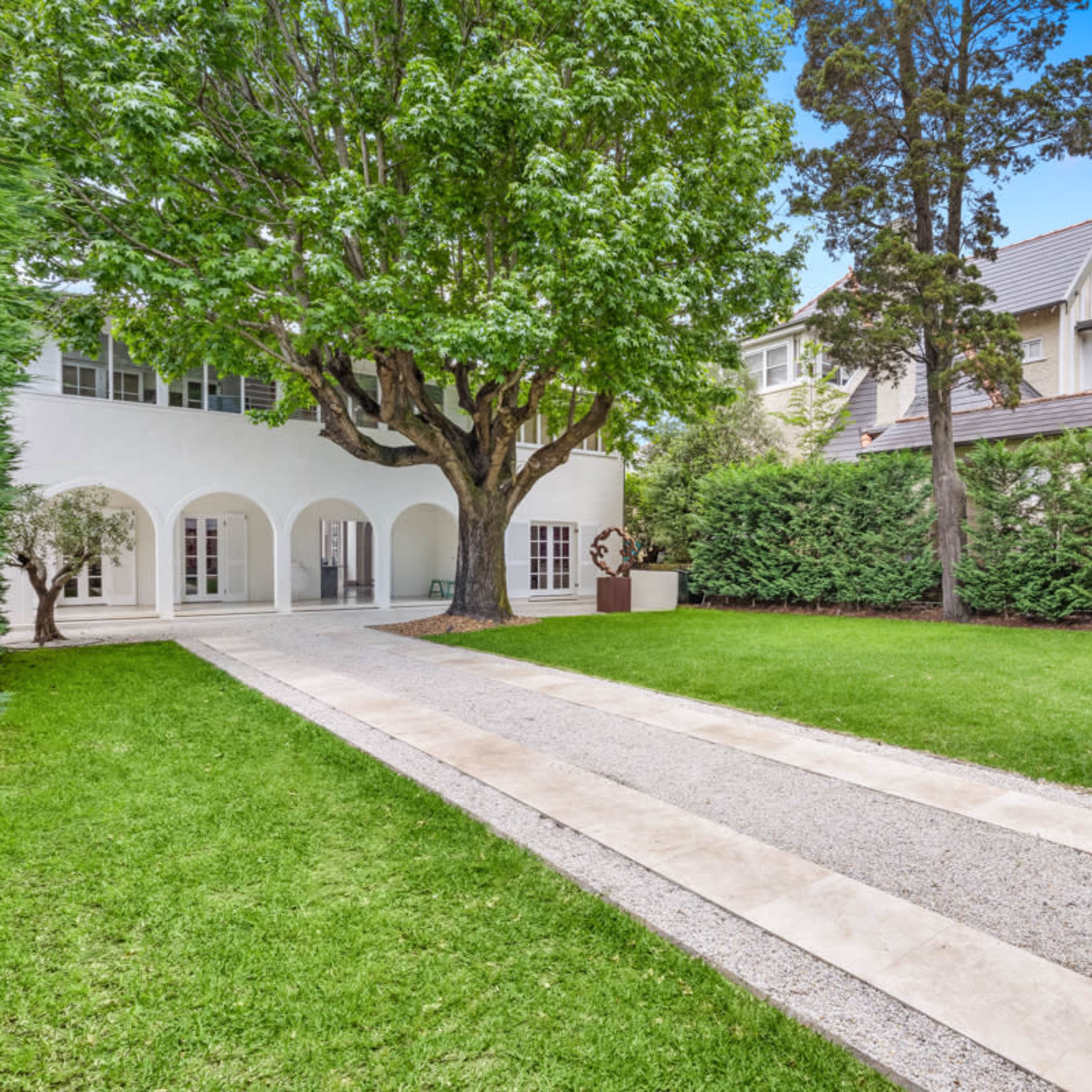 A gravel pathway leads to a white-walled house surrounded by manicured lawns and tall greenery.