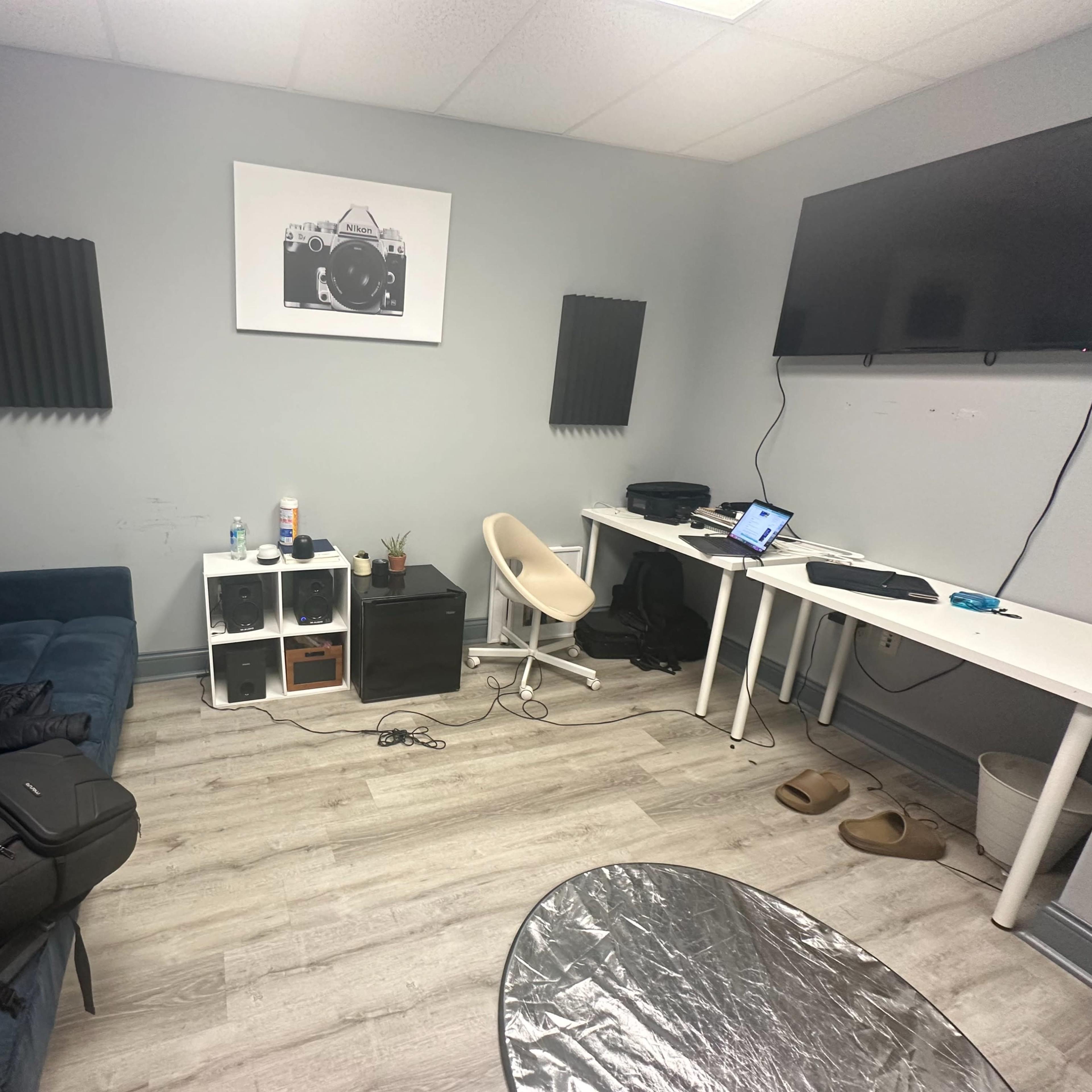 The room features a blue couch, a white desk with a computer, and organizers against a gray wall, with soundproofing panels on either side and a large TV.