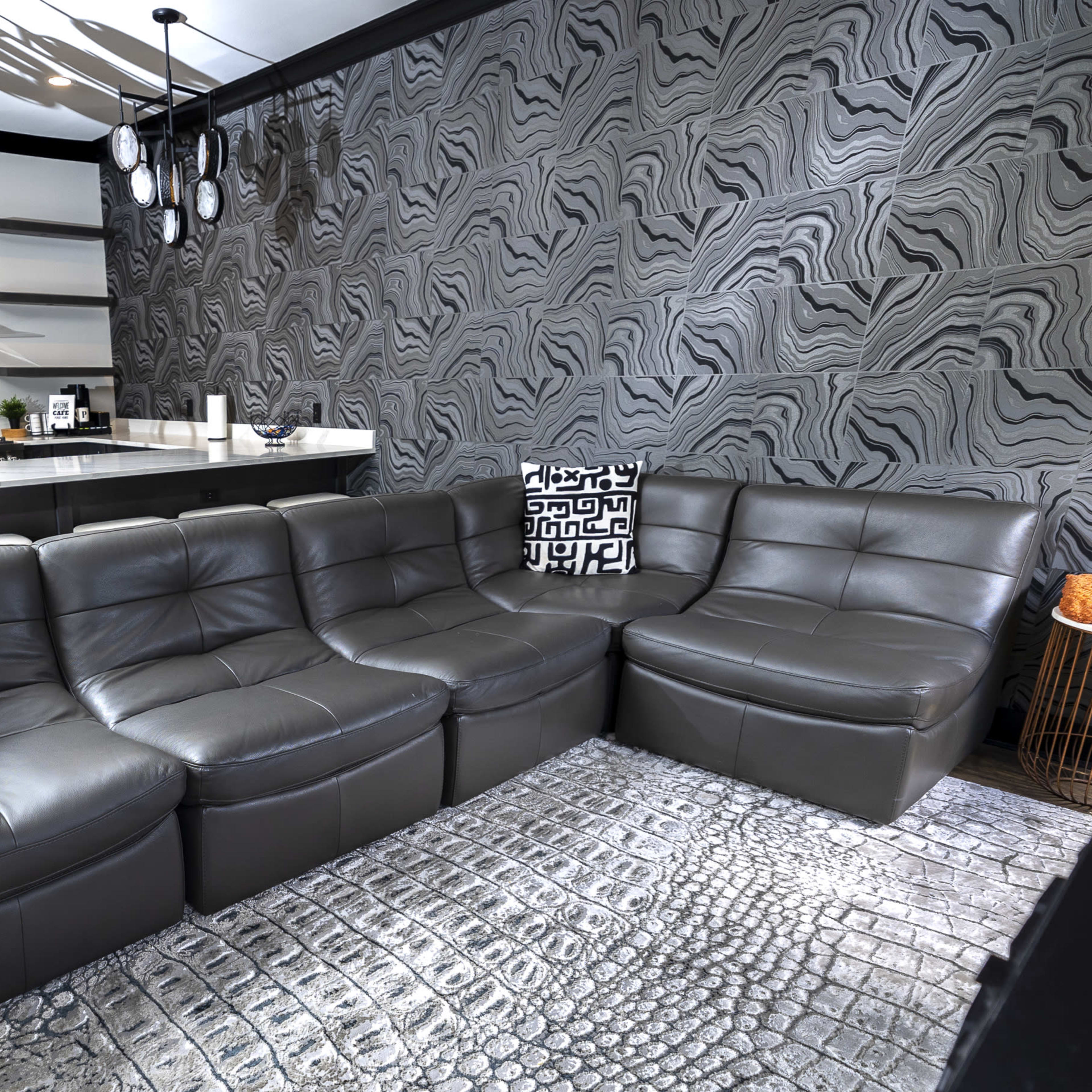 A modern living room with a dark gray leather sectional sofa, patterned throw pillows, and a patterned rug, alongside a sleek kitchen with a stainless steel oven and minimalist cabinetry.