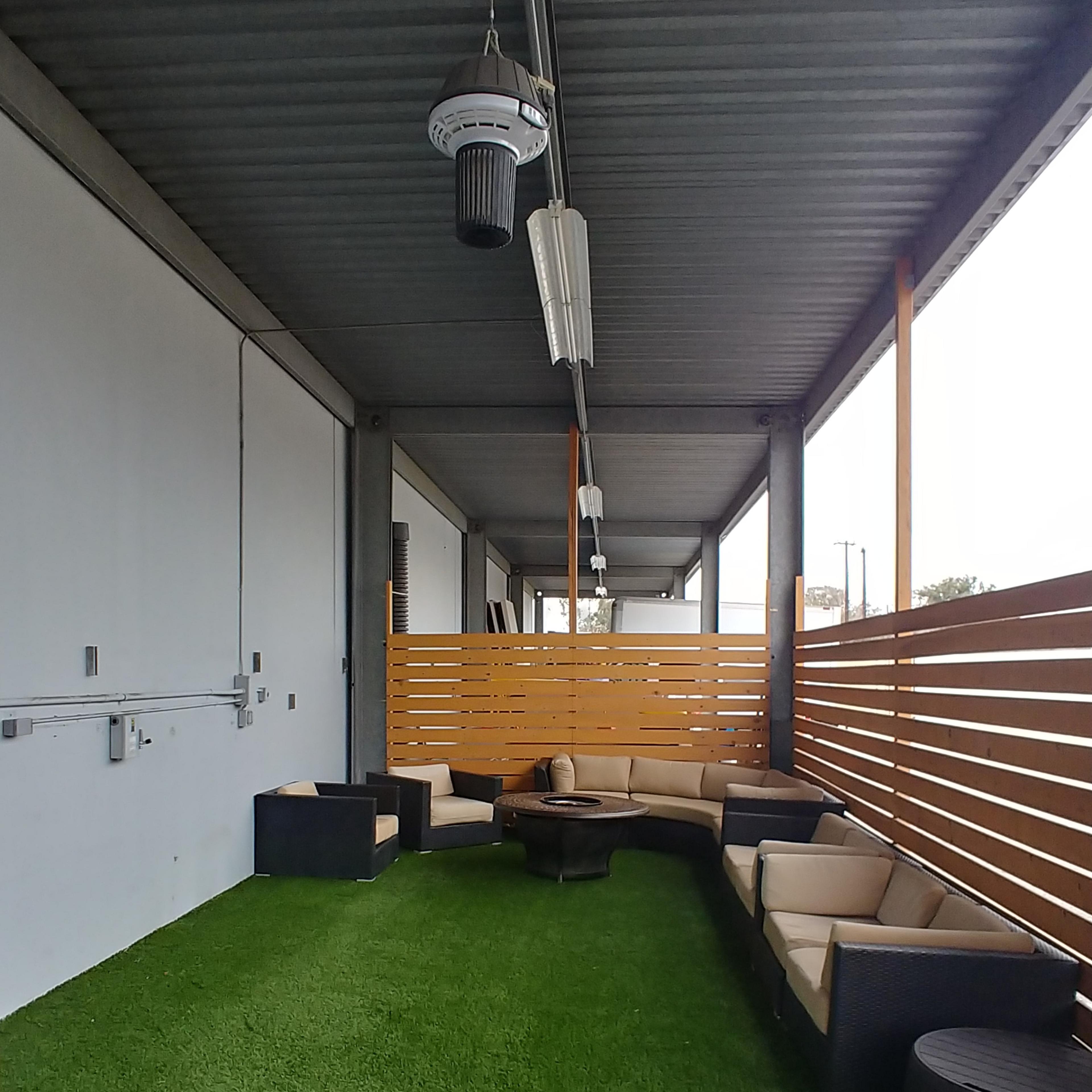A covered outdoor seating area with artificial grass, featuring a circular table and sectional couches along a wooden slatted partition.