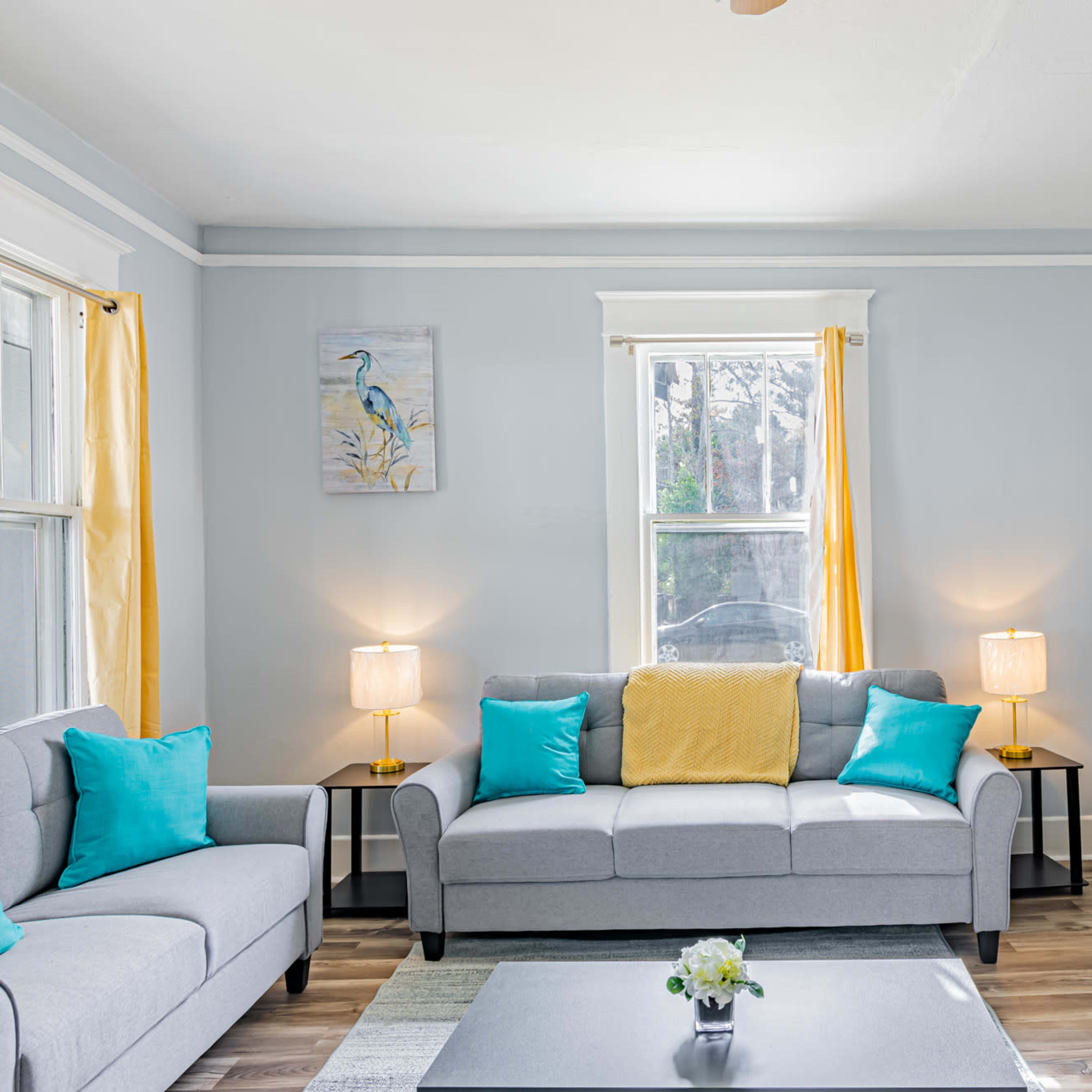 The living room features a gray sofa set with blue and yellow accent cushions, two lamps on side tables, and light-colored walls.