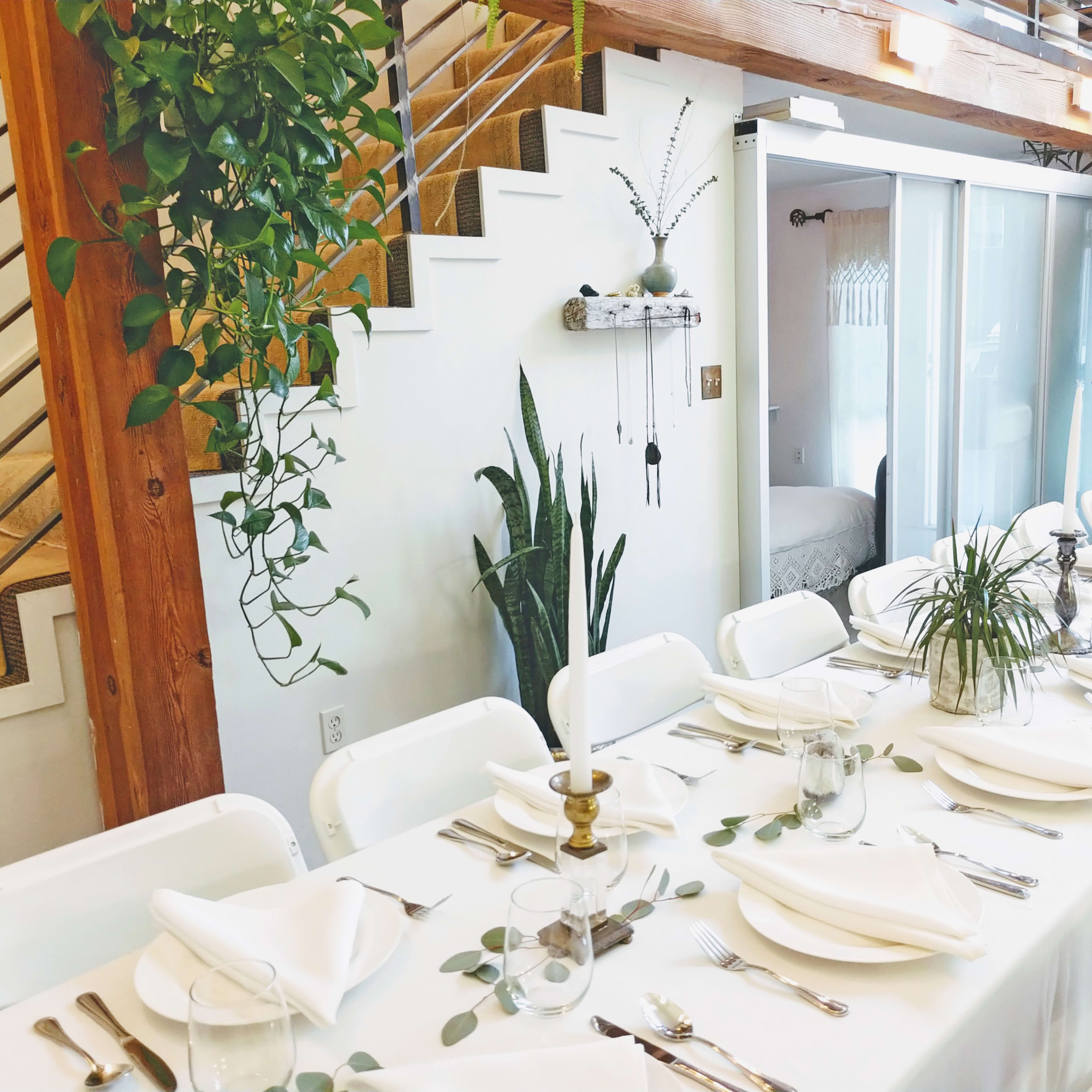 A long, neatly set dining table with white tablecloths and silverware is arranged under a staircase, surrounded by potted plants and a candle.