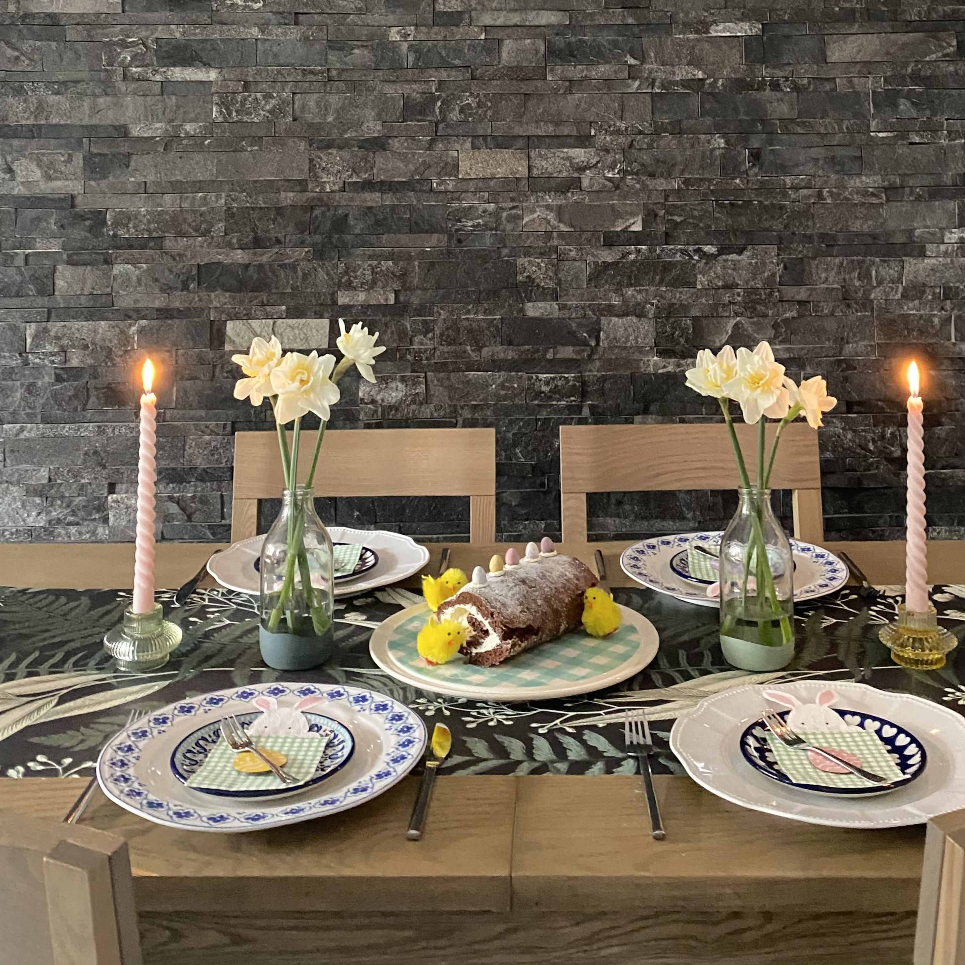 A dining table is set with plates, glasses, candles, and a decorated cake, surrounded by vases of flowers against a stone wall.
