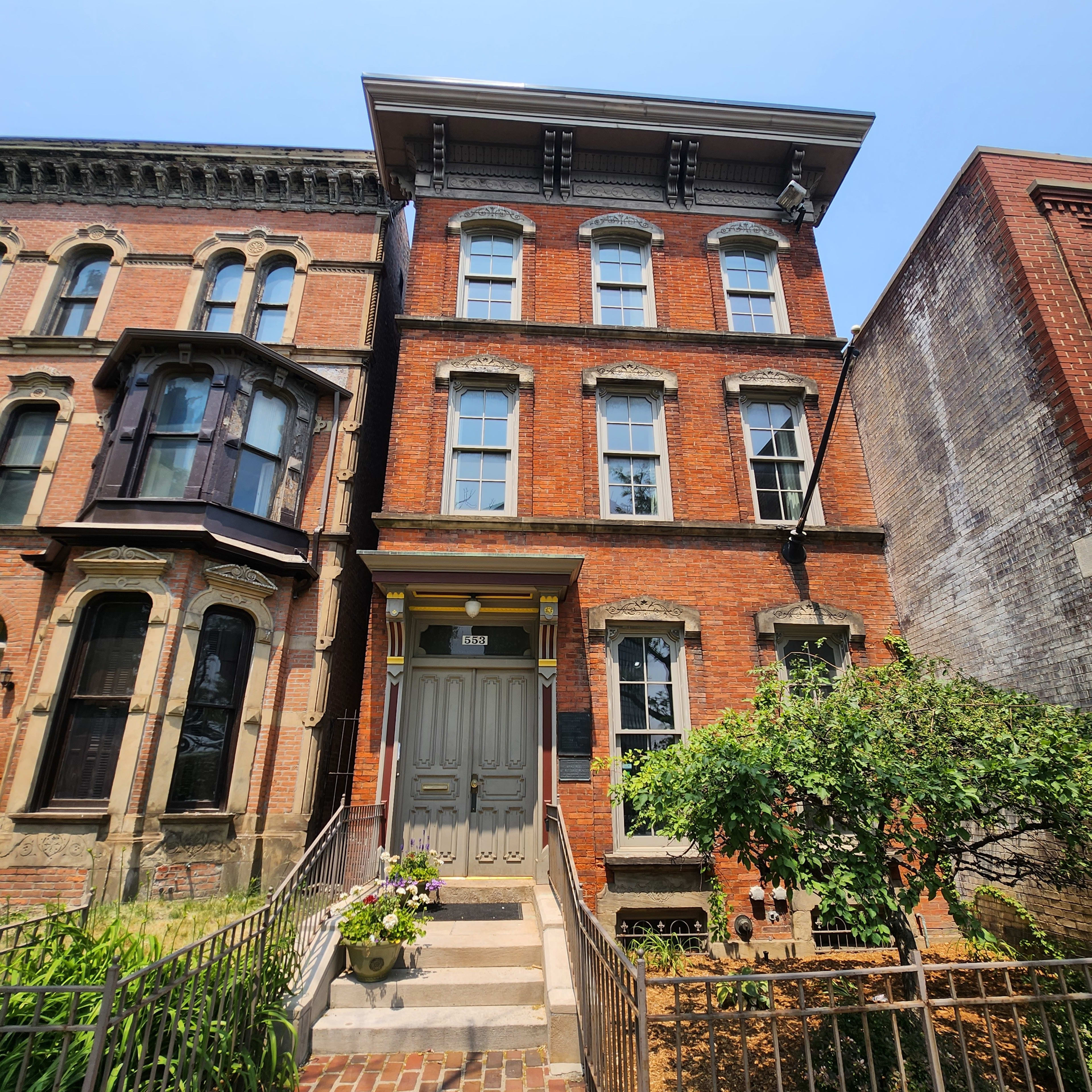 A three-story brick townhouse with large windows and a front staircase, flanked by a garden and neighboring buildings.