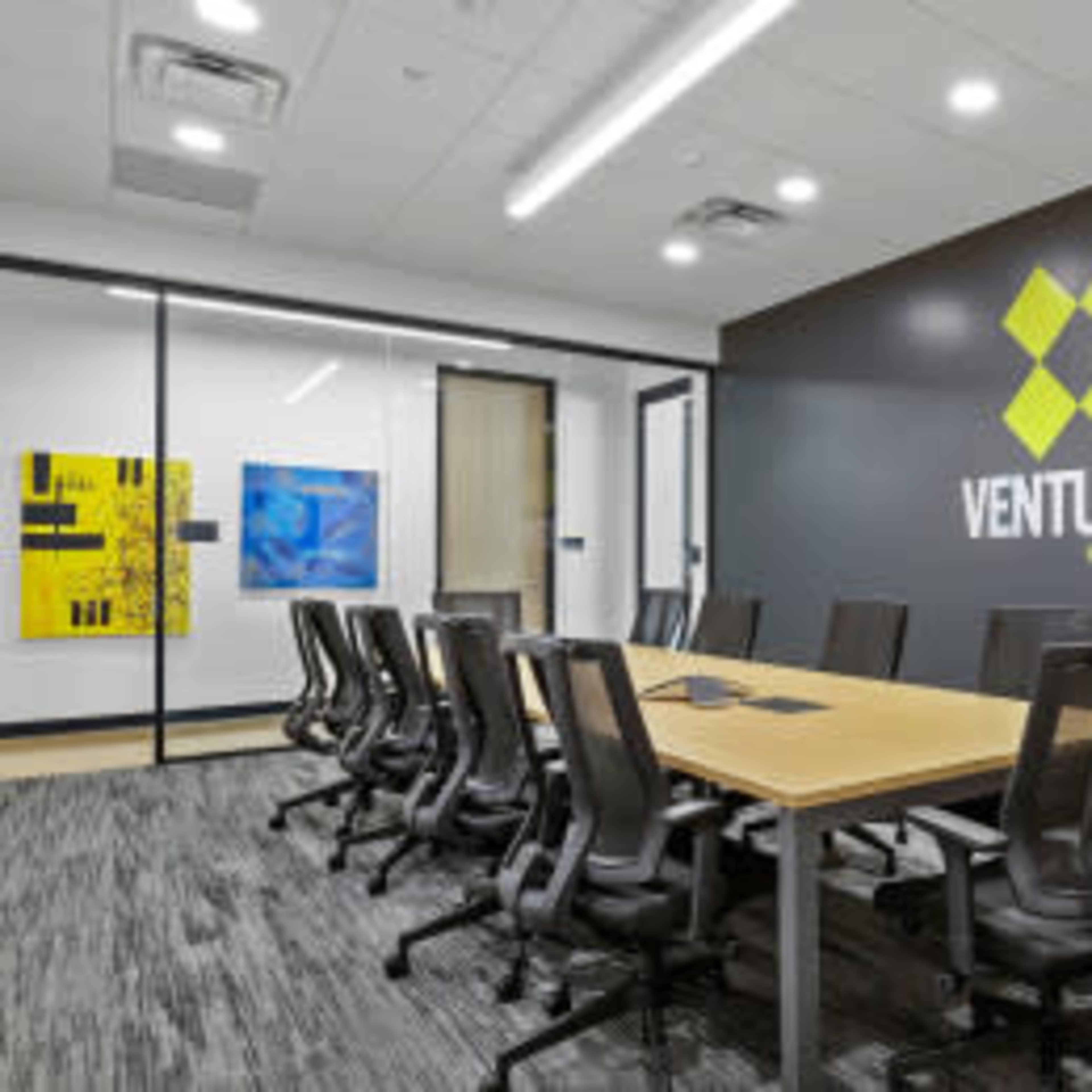 A modern conference room features a long wooden table surrounded by black chairs, with a large logo on the wall and colorful artwork displayed.