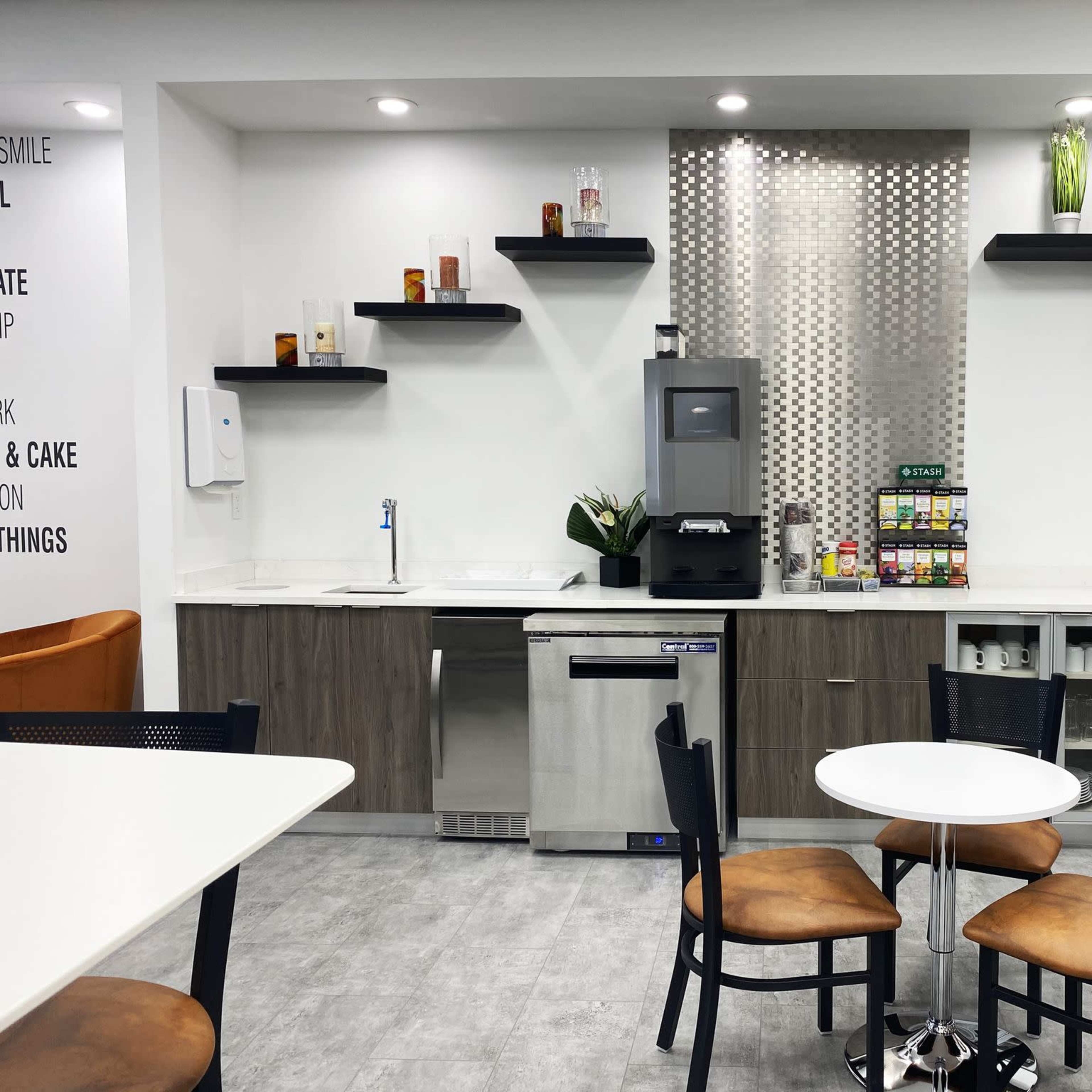 A modern kitchen area features a small round table with chairs, a sink, a refrigerator, shelves with various items, and a coffee station.