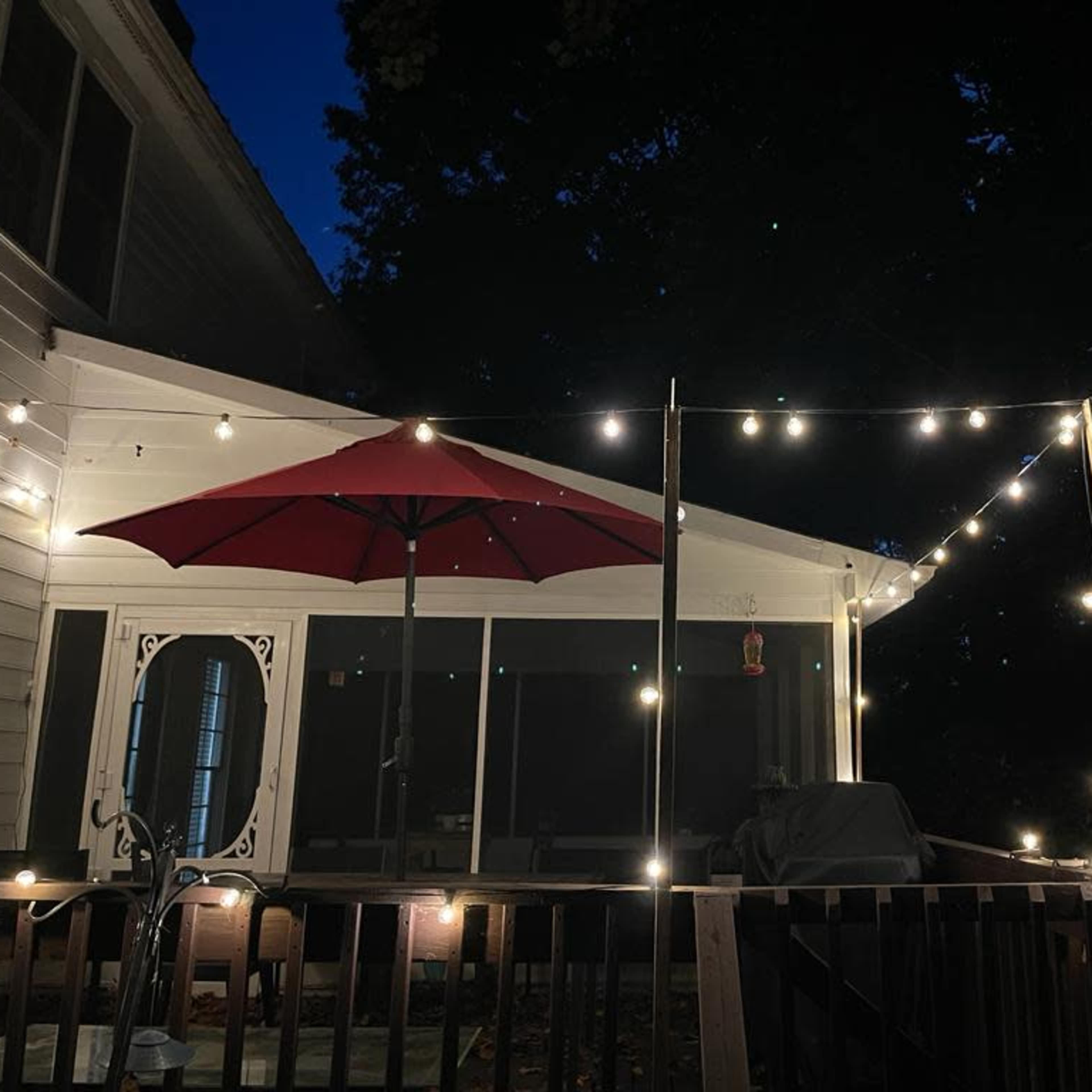 A deck illuminated by string lights, featuring an umbrella and a grill against a backdrop of trees at night.